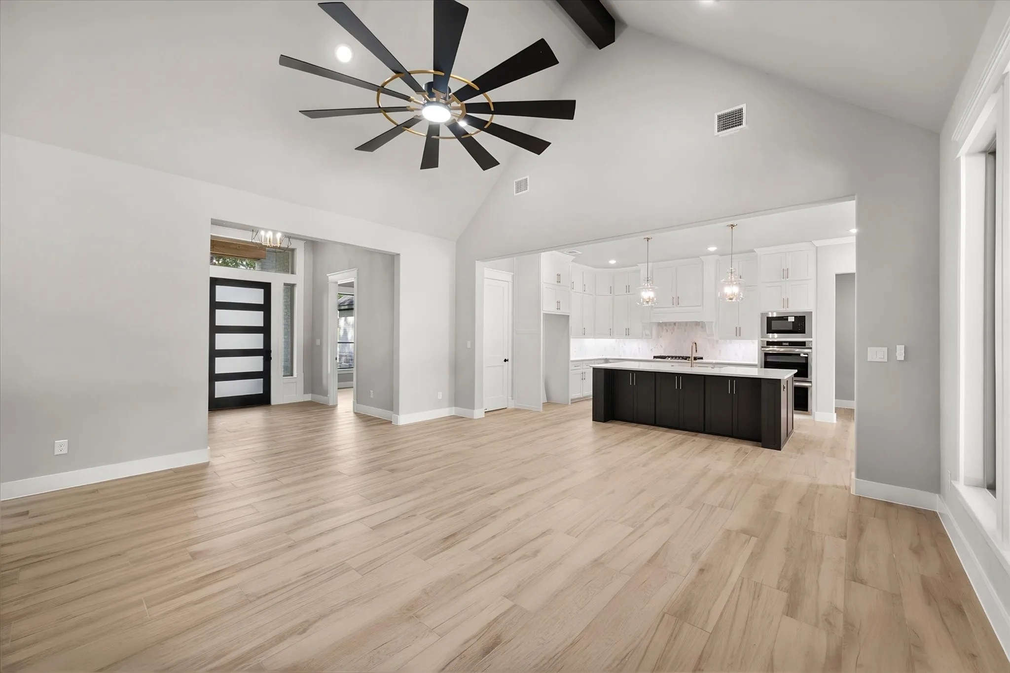 Open-concept living space featuring light wood-finish flooring throughout, a modern ceiling fan with integrated lighting, and seamless sightlines to a chef's kitchen with dark cabinetry, stainless steel appliances, and pendant lighting