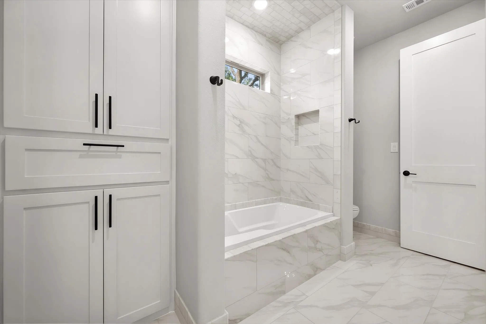 Built-in white cabinetry with black hardware, white tile flooring, white soaking tub, and white tiled shower surround with a recessed niche