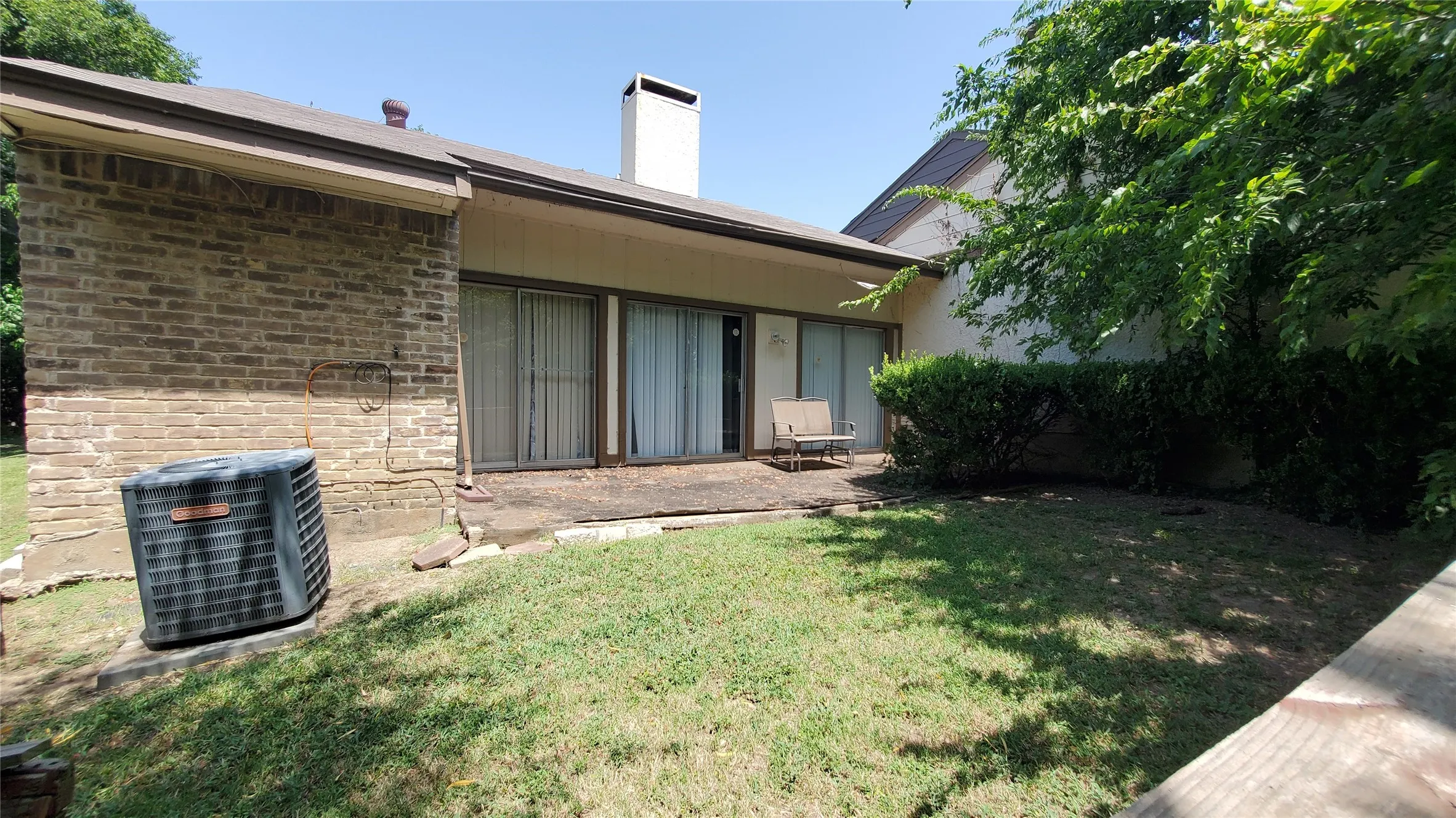 Rear exterior featuring brick siding, a concrete patio, two sliding glass doors, a white chimney, and established shrubbery