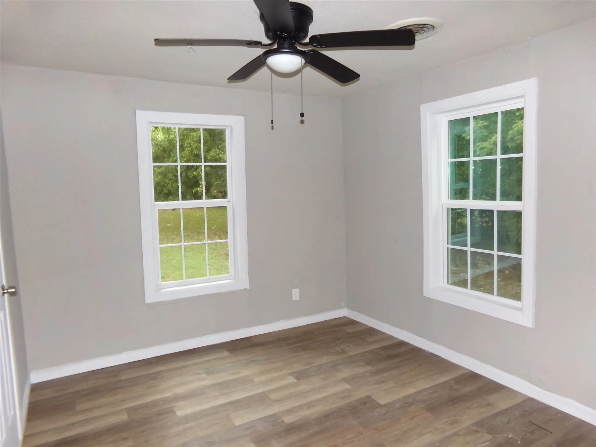 Room featuring wood-finish flooring, two double-hung windows with white trim, a ceiling fan with light fixture, and light-toned wall paint