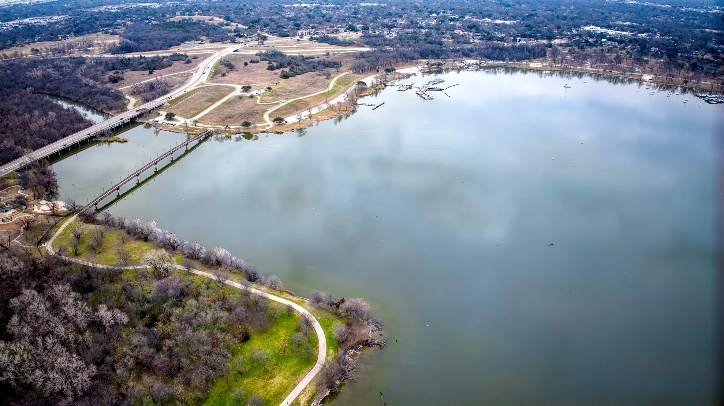 White Rock Lake featuring a marina with multiple boat docks, a bridge spanning across the water, surrounding wooded areas, and paved pedestrian paths along the shoreline