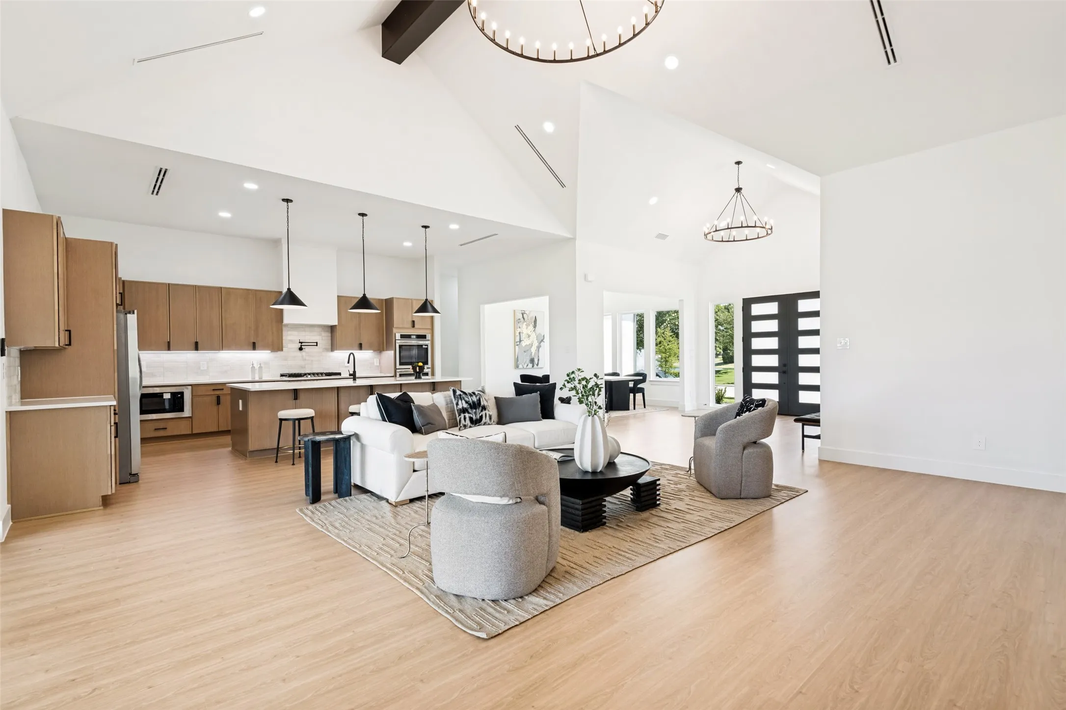 Spacious open-concept living area featuring a vaulted ceiling with exposed beam, light wood-finish flooring, and a contemporary kitchen with light wood-tone cabinetry, stainless steel appliances, and a central island with pendant lighting
