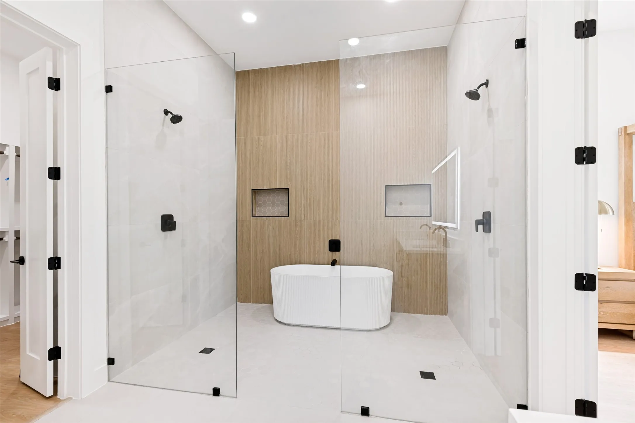 Contemporary bathroom featuring a freestanding soaking tub, dual glass-enclosed shower areas with matte black fixtures, recessed lighting, and contrasting light and wood-finish wall tiling