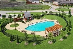 Resort-style pool area featuring a freeform swimming pool, extensive concrete decking, a wooden pergola, a rustic-style poolside building, and surrounding green space with a black metal fence