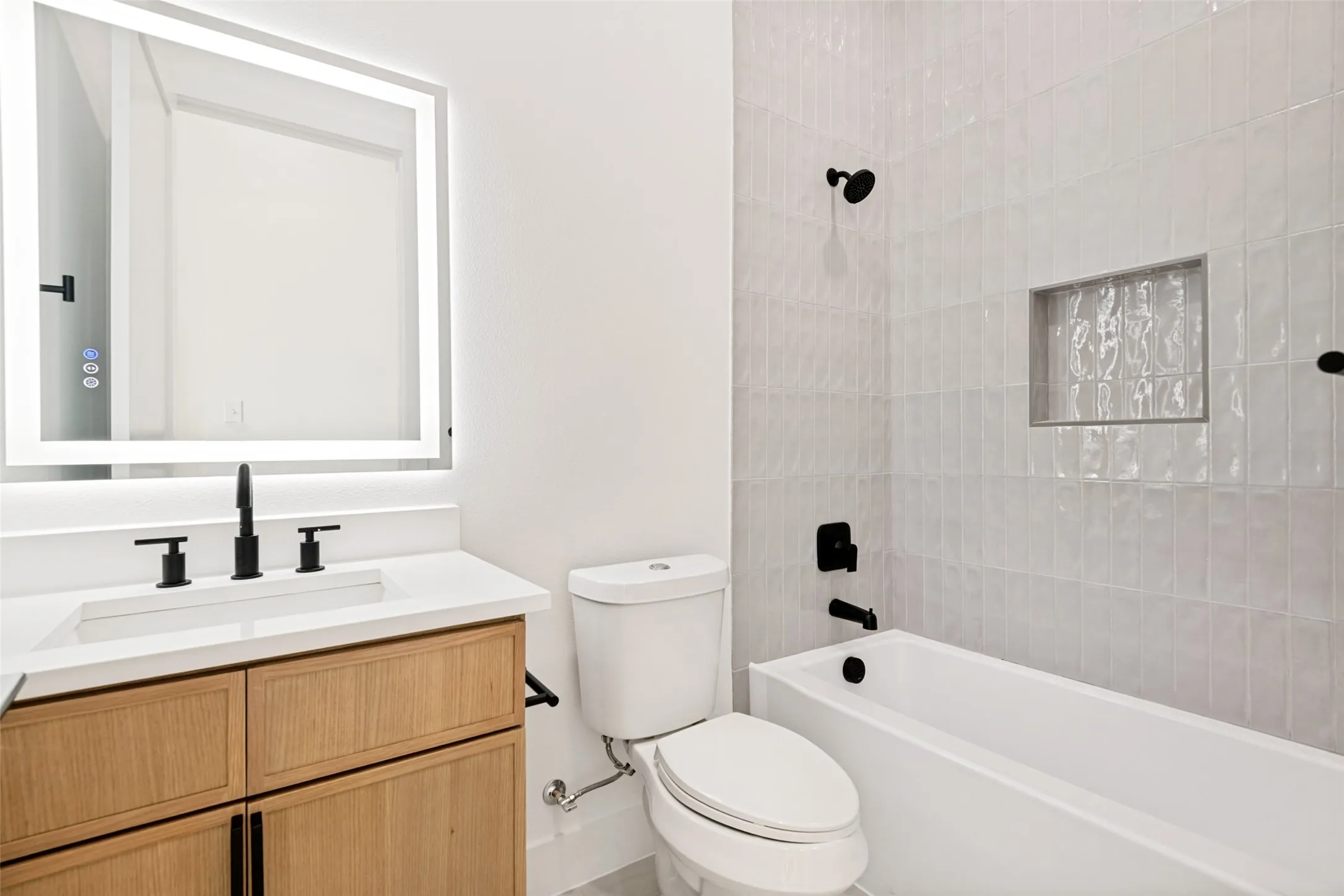 White bathroom featuring a light wood-finish vanity with a white countertop and integrated sink, matte black fixtures, an illuminated mirror, and a white subway tile surround for the tub and shower