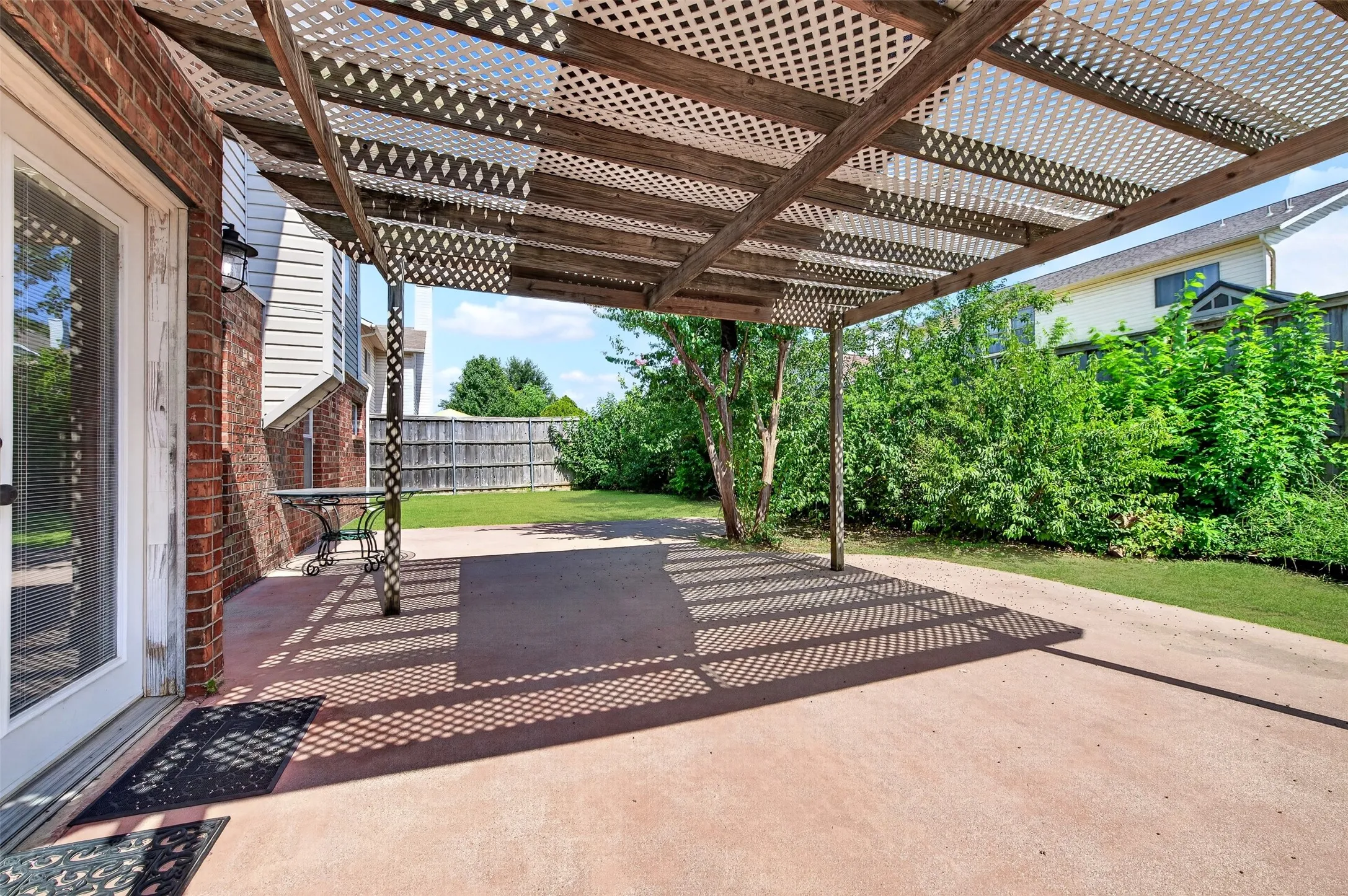 Expansive patio featuring a lattice pergola, direct access from the property, mature perimeter greenery, and a wooden privacy fence