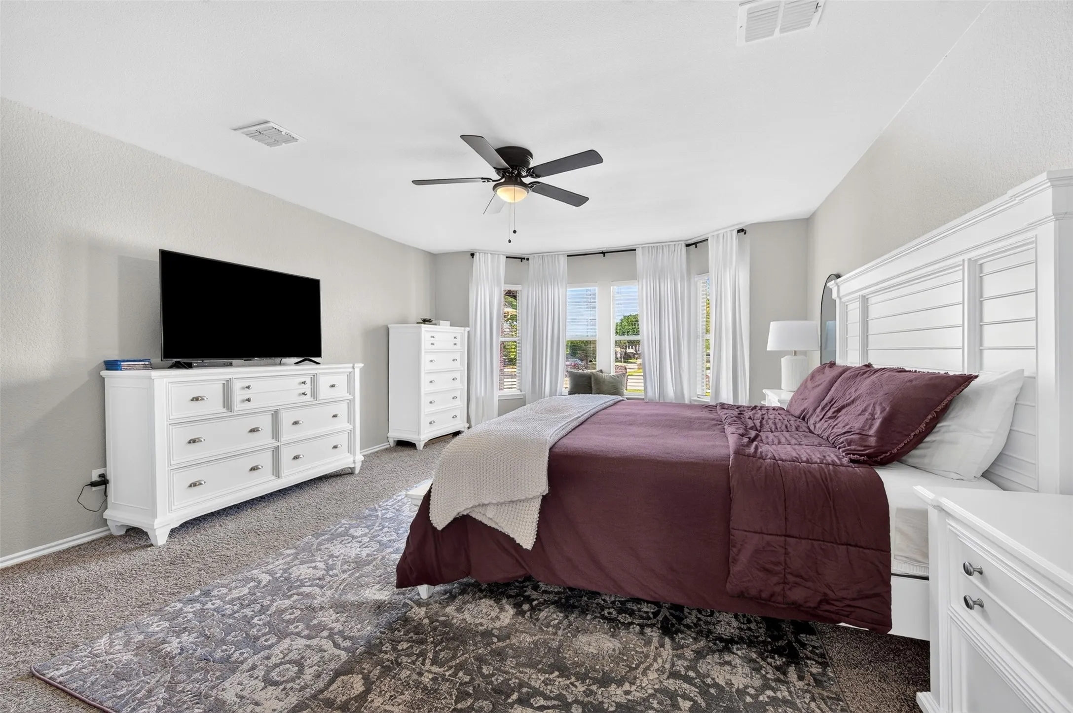 Spacious bedroom featuring light gray walls, carpet flooring, a ceiling fan, and a prominent bay window with white curtains