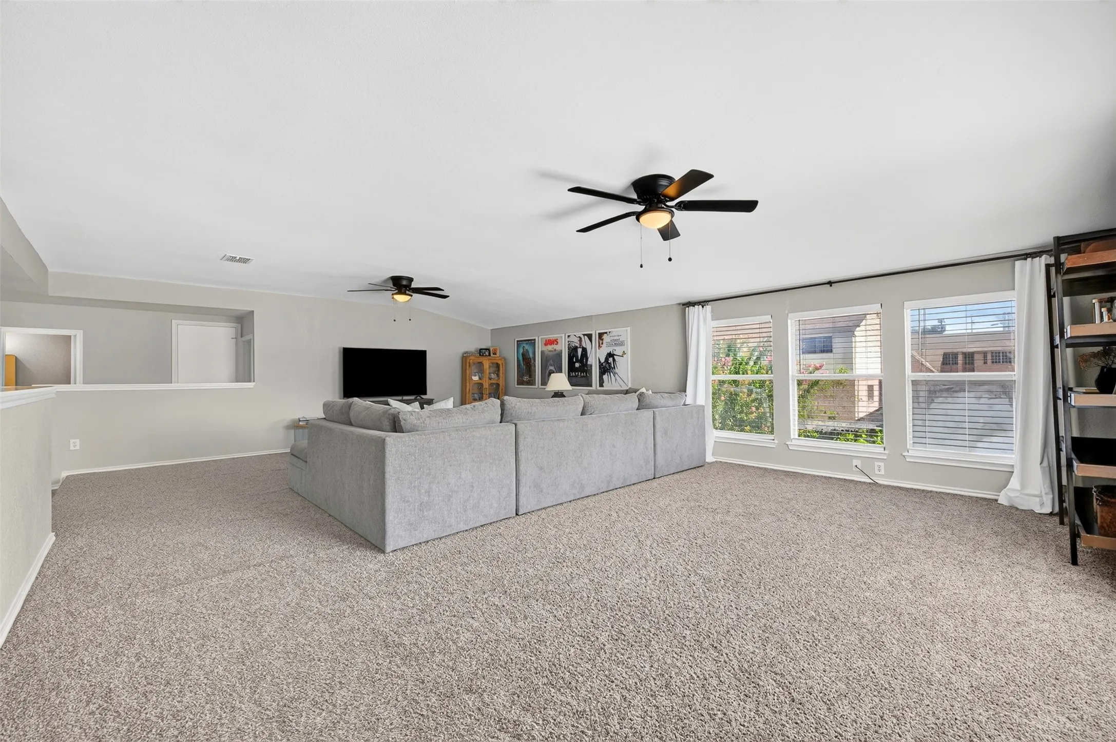 Spacious carpeted room featuring multiple windows with blinds, two ceiling fans with lighting, and a partial wall with a cutout opening
