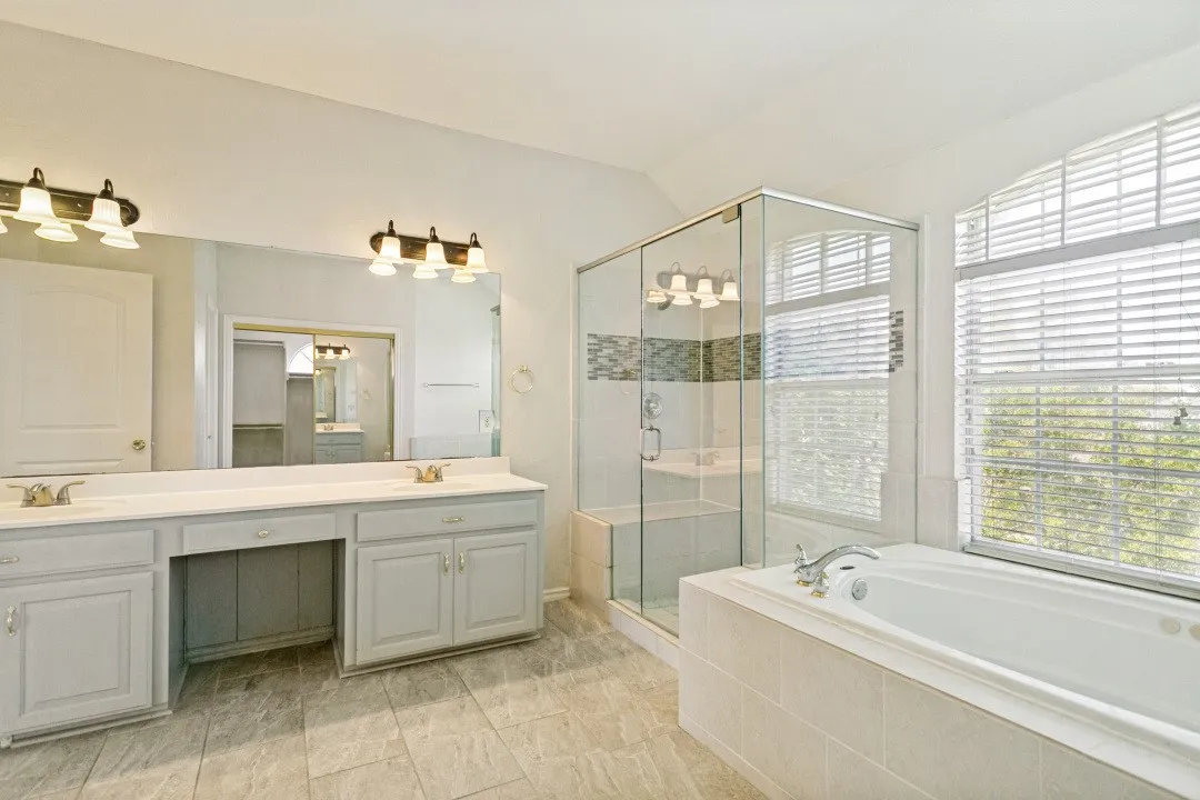 Expansive bathroom featuring a tiled floor, dual vanity with light cabinetry, large mirror, glass-enclosed shower with tiled accents, and a built-in soaking tub beneath a window