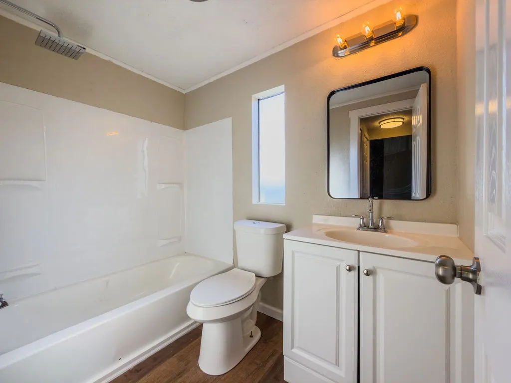 Bathroom featuring a tub/shower combination with a square rainfall showerhead, a white vanity with panel doors and silver-tone hardware, a rectangular mirror, and wood-finish flooring