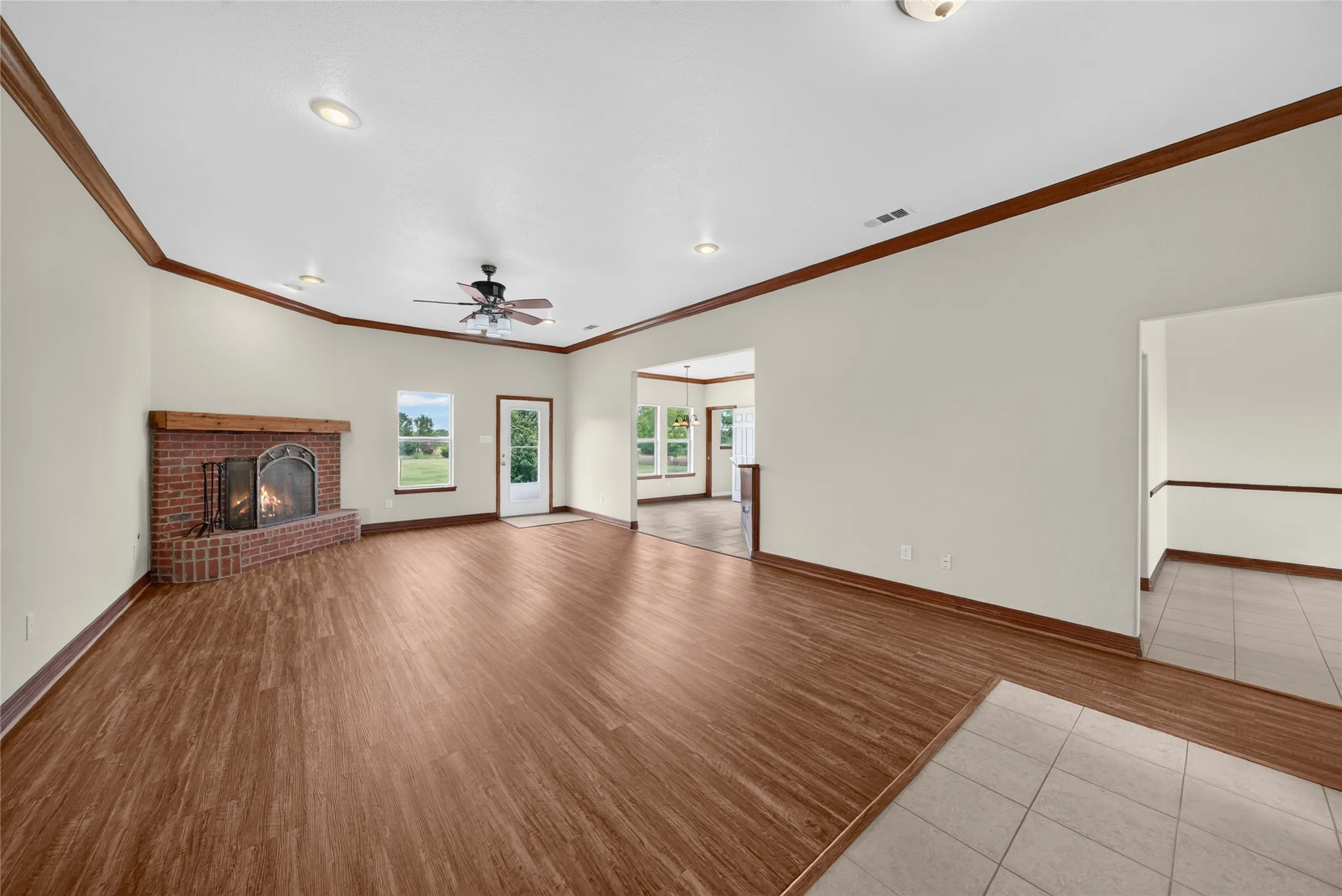 Spacious living area featuring wood-finish flooring, a brick fireplace with a wood mantle, white walls, and dark wood crown molding