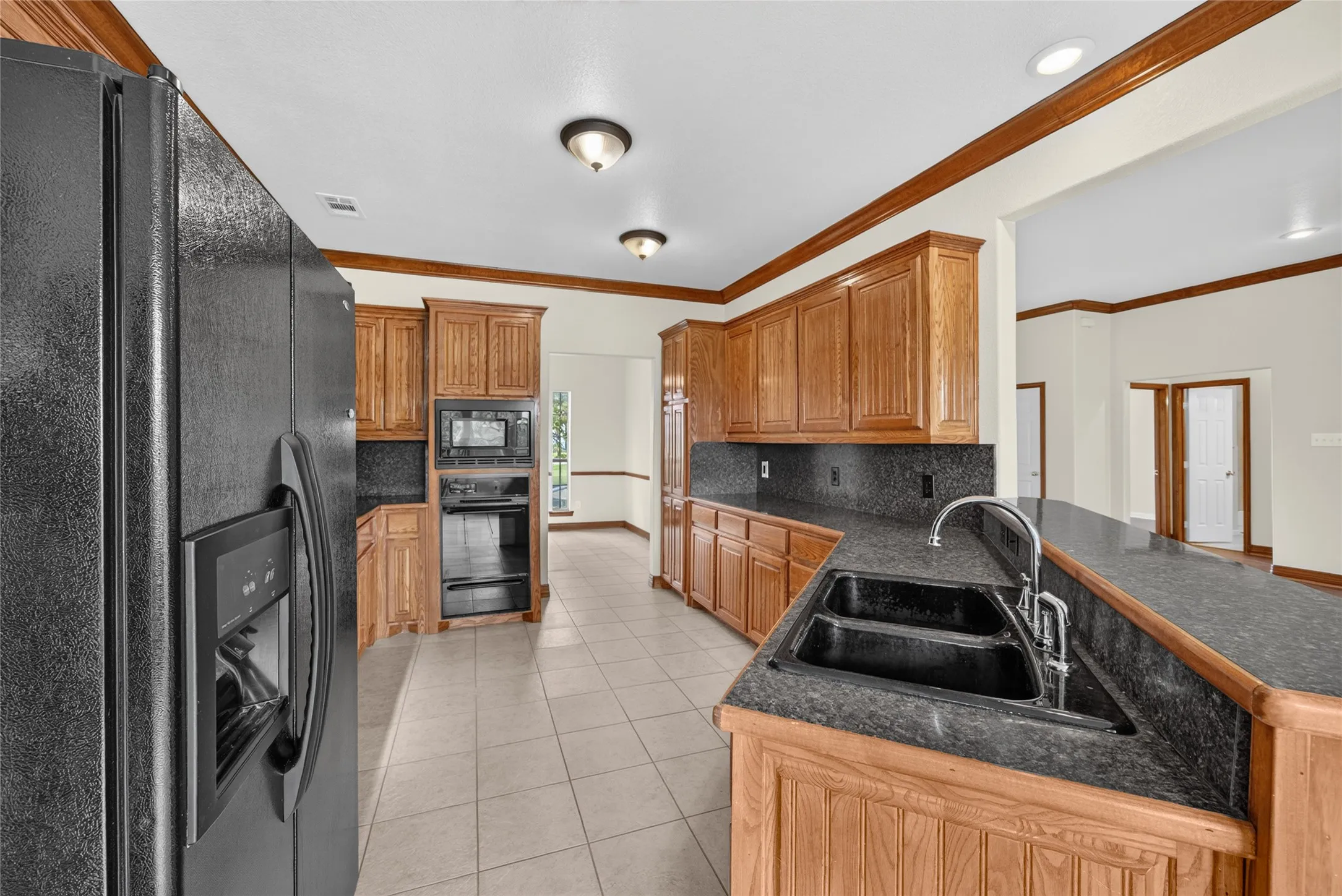 Spacious kitchen featuring extensive wood cabinetry, dark countertops, black double basin sink with chrome faucet, black built-in oven and microwave, and tiled flooring