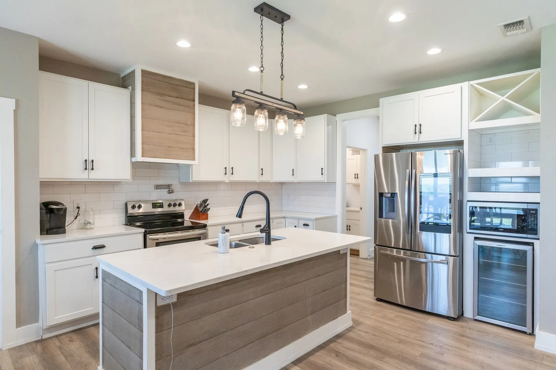 Modern kitchen featuring white cabinetry, light wood-finish flooring, a central island with a light countertop, stainless steel appliances, and a contemporary linear chandelier