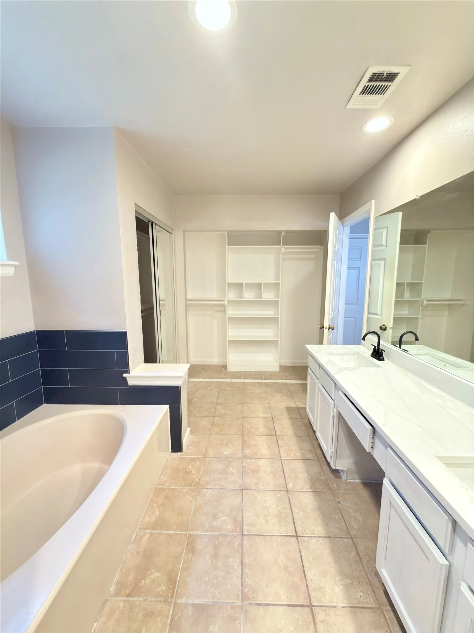 Spacious bathroom featuring a built-in soaking tub with dark blue tile surround, a double vanity with white cabinetry and integrated sinks, and ample built-in shelving