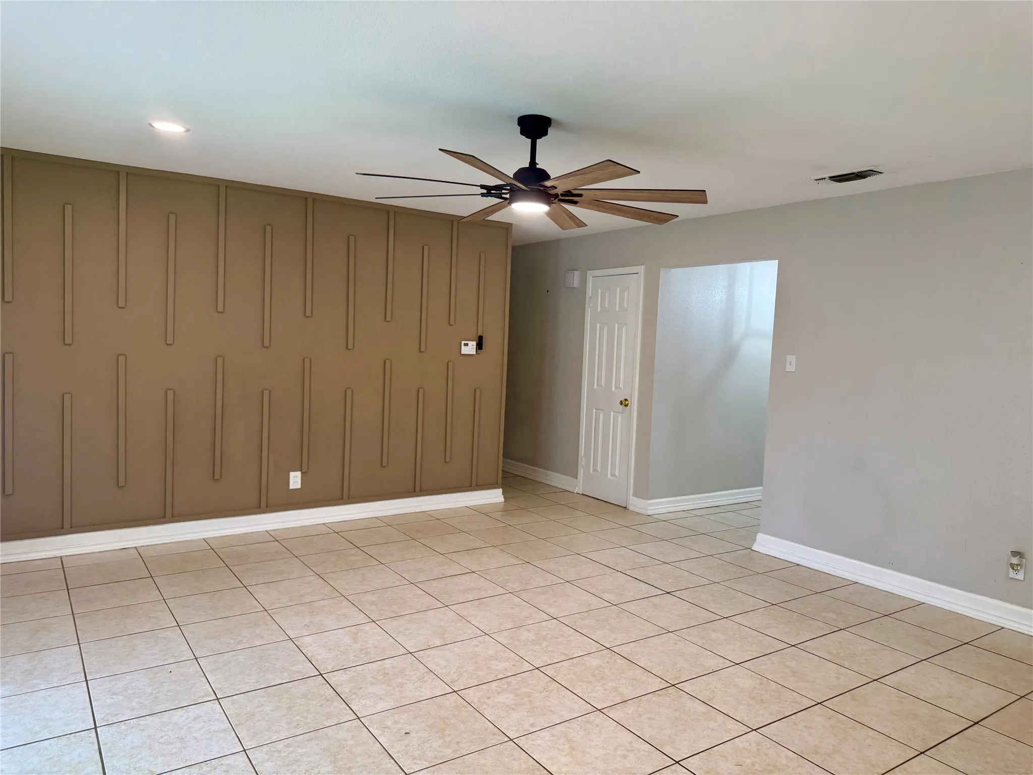 Spacious room featuring neutral-toned tile flooring, a decorative accent wall with vertical paneling, and a modern ceiling fan with wood-look blades