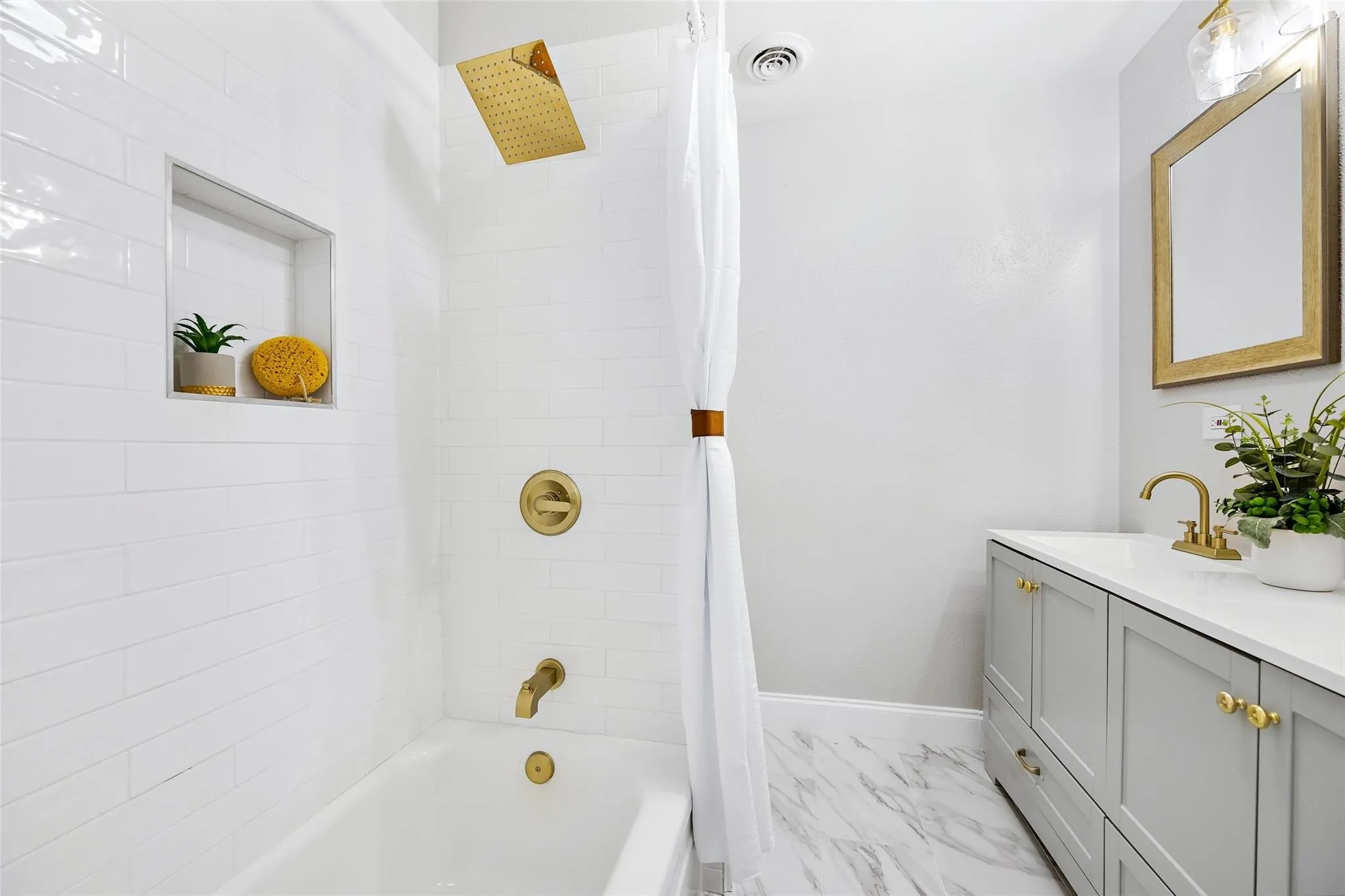 Bathroom featuring a white subway tile shower surround with a built-in niche, gold-tone fixtures, a white soaking tub, a gray vanity with a white countertop, and marble-look floor tiling