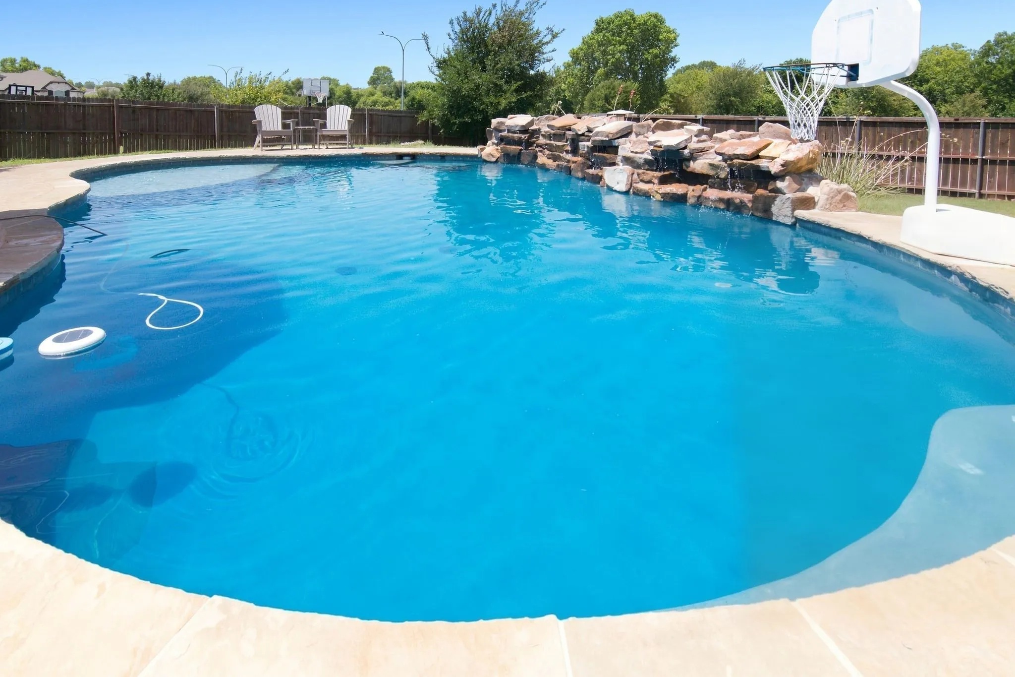 Curved swimming pool with natural stone waterfall feature, surrounding concrete decking, and a privacy fence