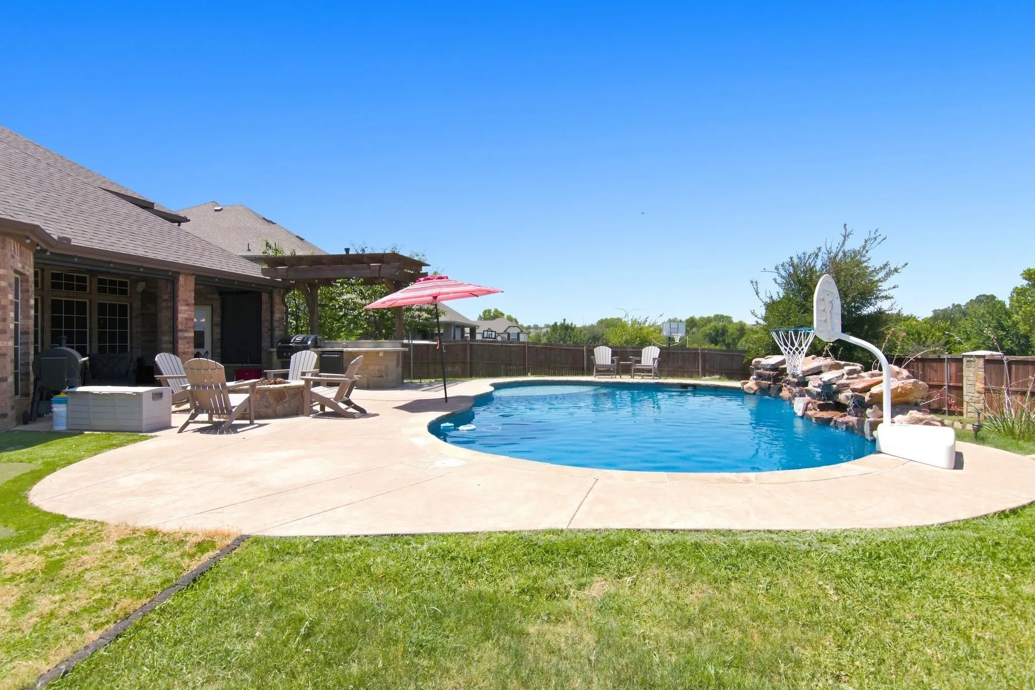 Resort-style backyard oasis featuring a freeform saltwater swimming pool with integrated spa, expansive concrete decking, mature landscaping, and a brick home with covered patio and pergola