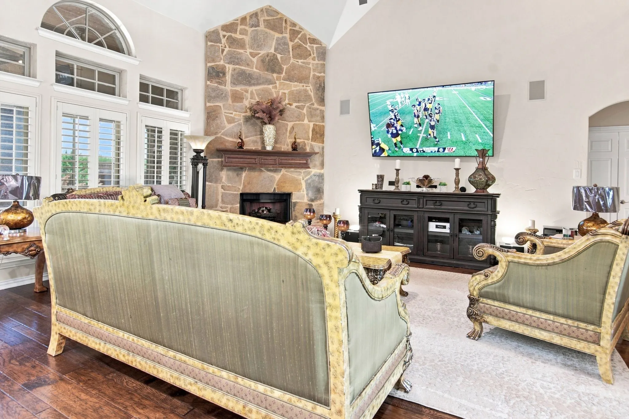 Vaulted ceiling living room featuring a floor-to-ceiling stone fireplace with a wood mantle, hardwood flooring, and multiple windows with plantation shutters