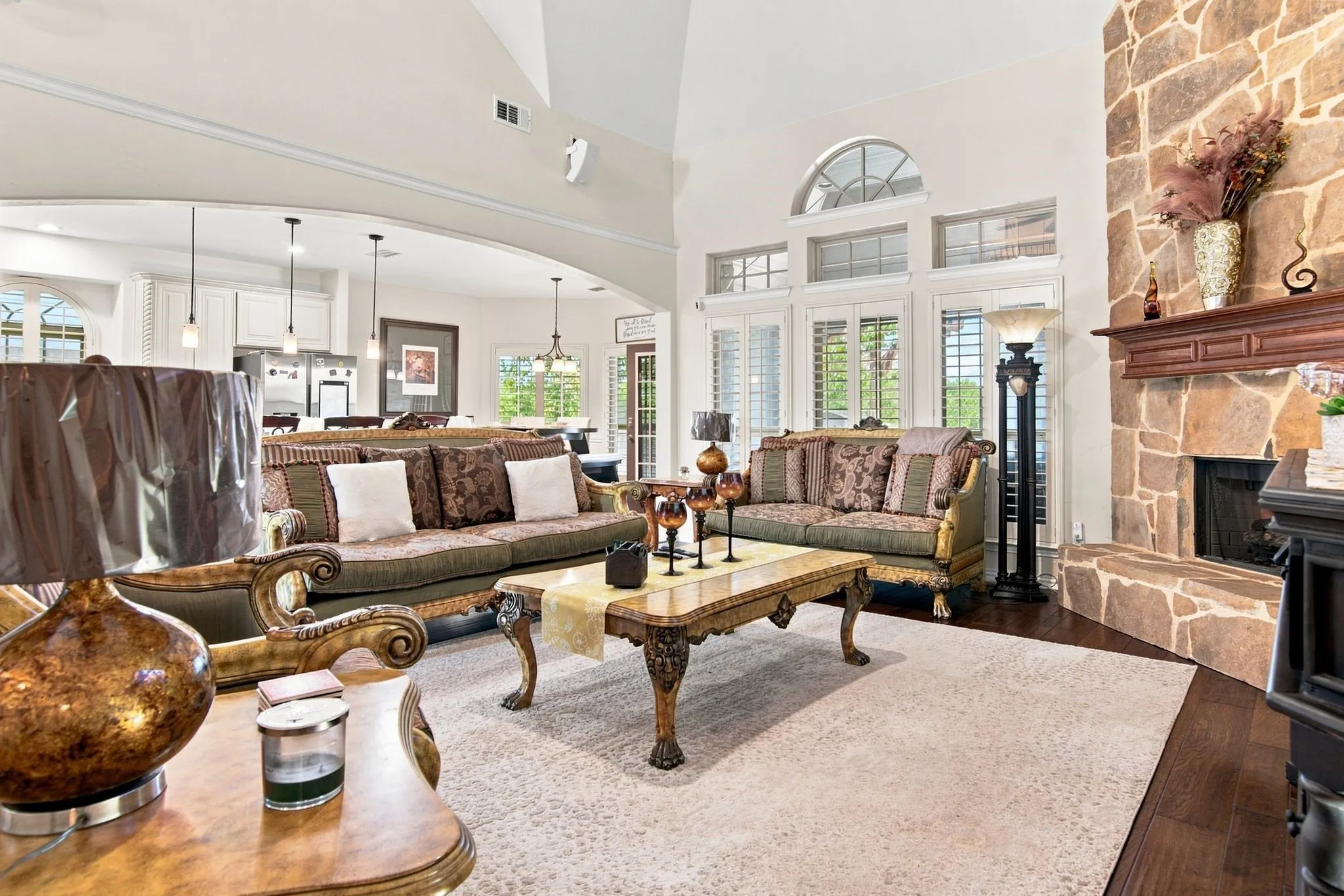 Spacious living area featuring a floor-to-ceiling stone fireplace, high ceilings, wood-finish flooring, and white plantation shutters