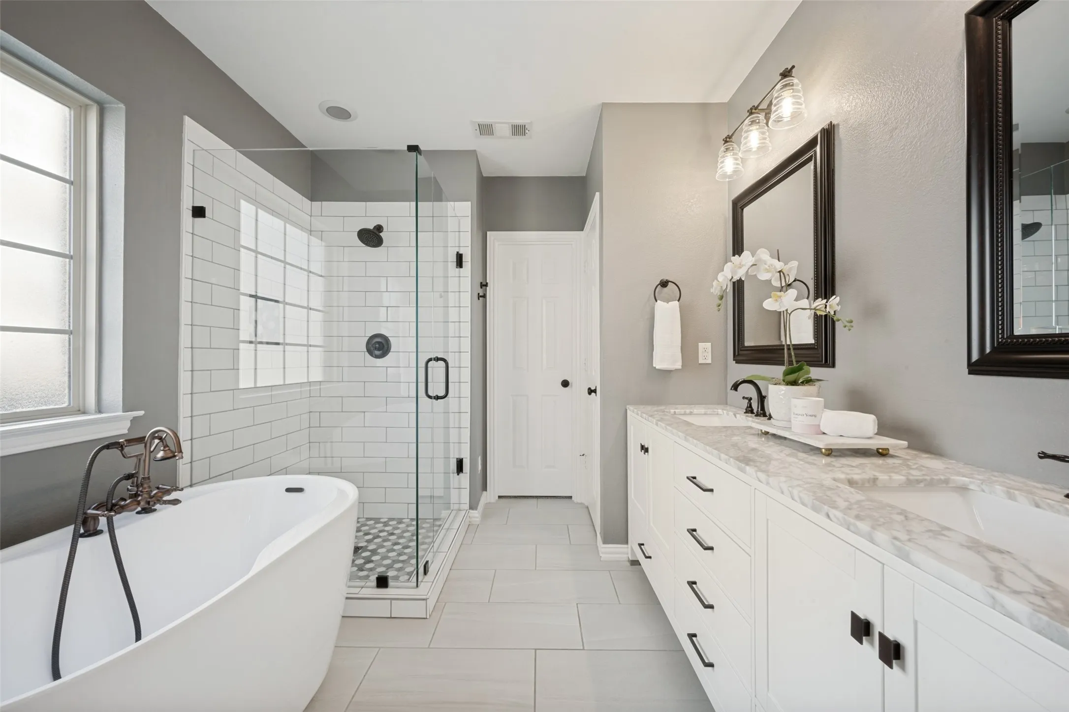 Step into a spa-inspired primary bath designed for both luxury and everyday comfort. This beautifully appointed retreat features a freestanding soaking slipper tub, a spacious frameless glass walk-in shower an expansive dual-sink vanity topped with quartz.  Soft gray walls, crisp white cabinetry, matte black fixtures, and stylish lighting create a timeless, sophisticated aesthetic, while abundant natural light enhances the bright and airy feel. Thoughtfully designed with generous storage and high-end finishes throughout, this serene space offers the perfect place to unwind.