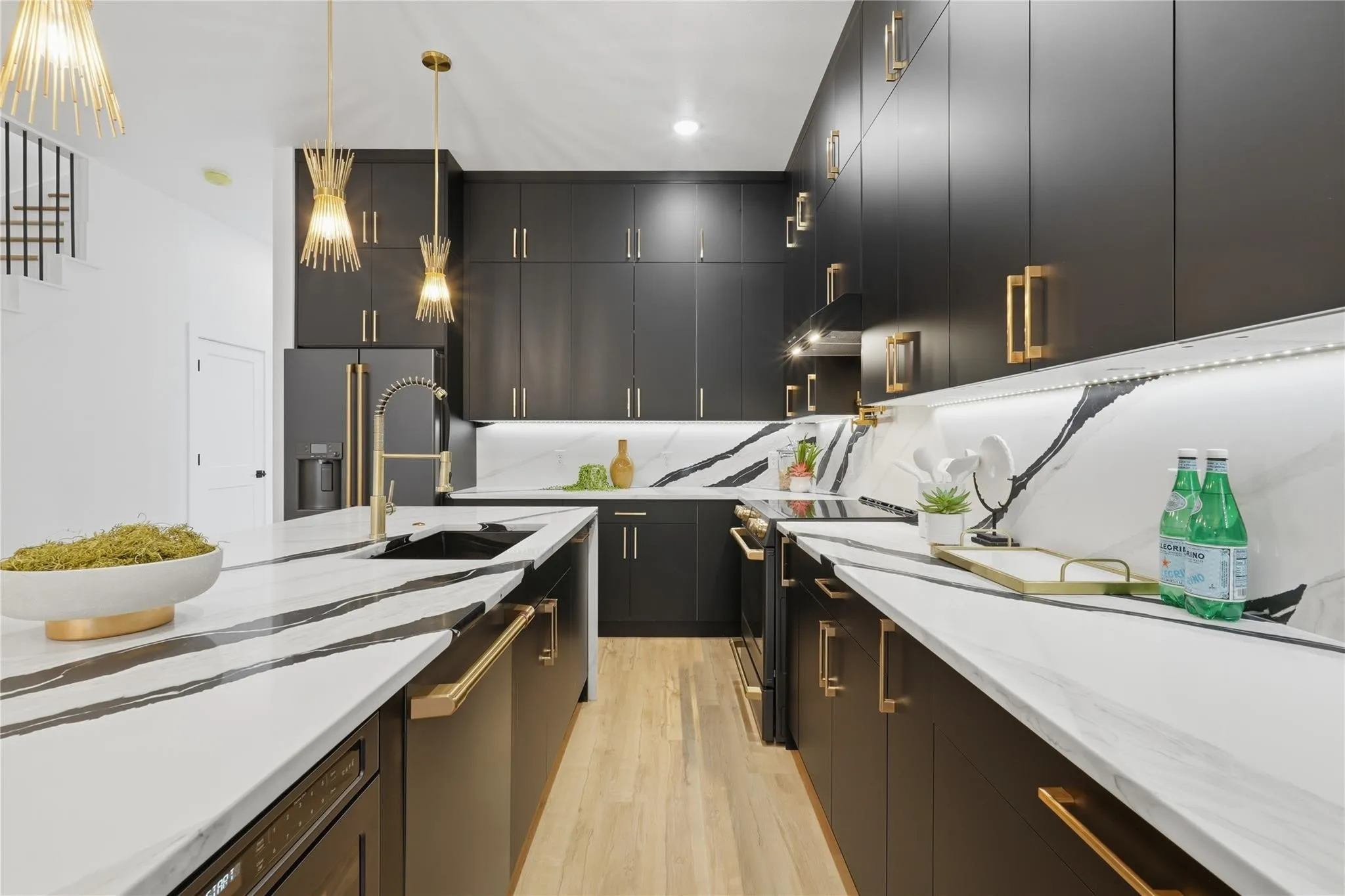 Modern kitchen featuring black cabinetry with gold hardware, white countertops with prominent black veining, integrated appliances, and light wood-finish flooring