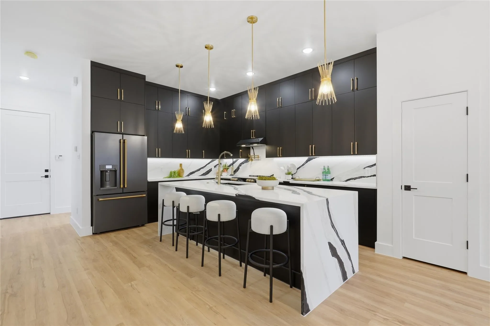 Contemporary kitchen featuring dark flat-panel cabinetry with gold-tone hardware, a large island with waterfall-edge countertop, integrated appliances, and light wood-finish flooring