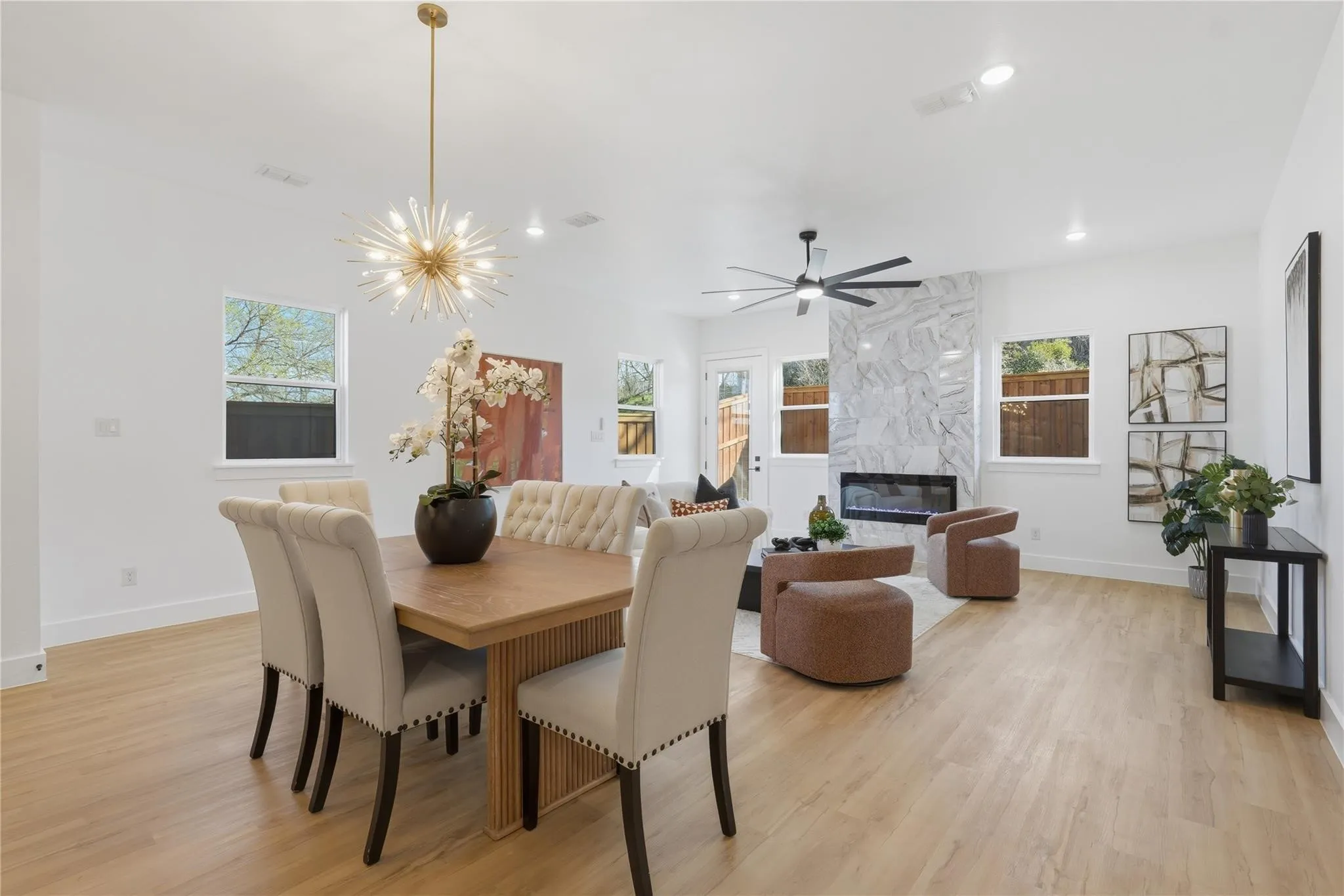 Open-concept living space featuring light wood-finish flooring, a modern fireplace with a light-toned stone surround, a contemporary ceiling fan, recessed lighting, and multiple windows providing natural light