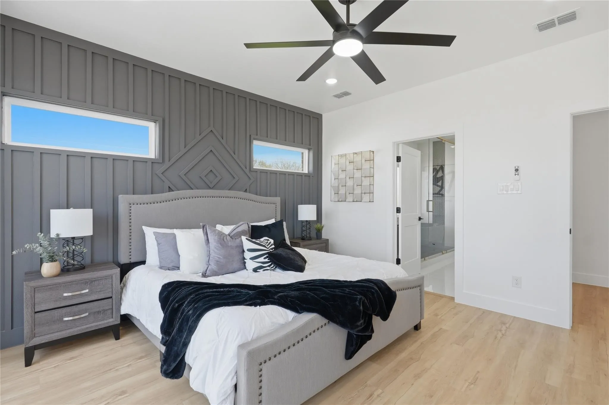 Bedroom featuring an accent wall with vertical paneling and geometric relief, light wood-finish flooring, recessed lighting, a ceiling fan with integrated light, and an entry to an en-suite with a glass-enclosed shower