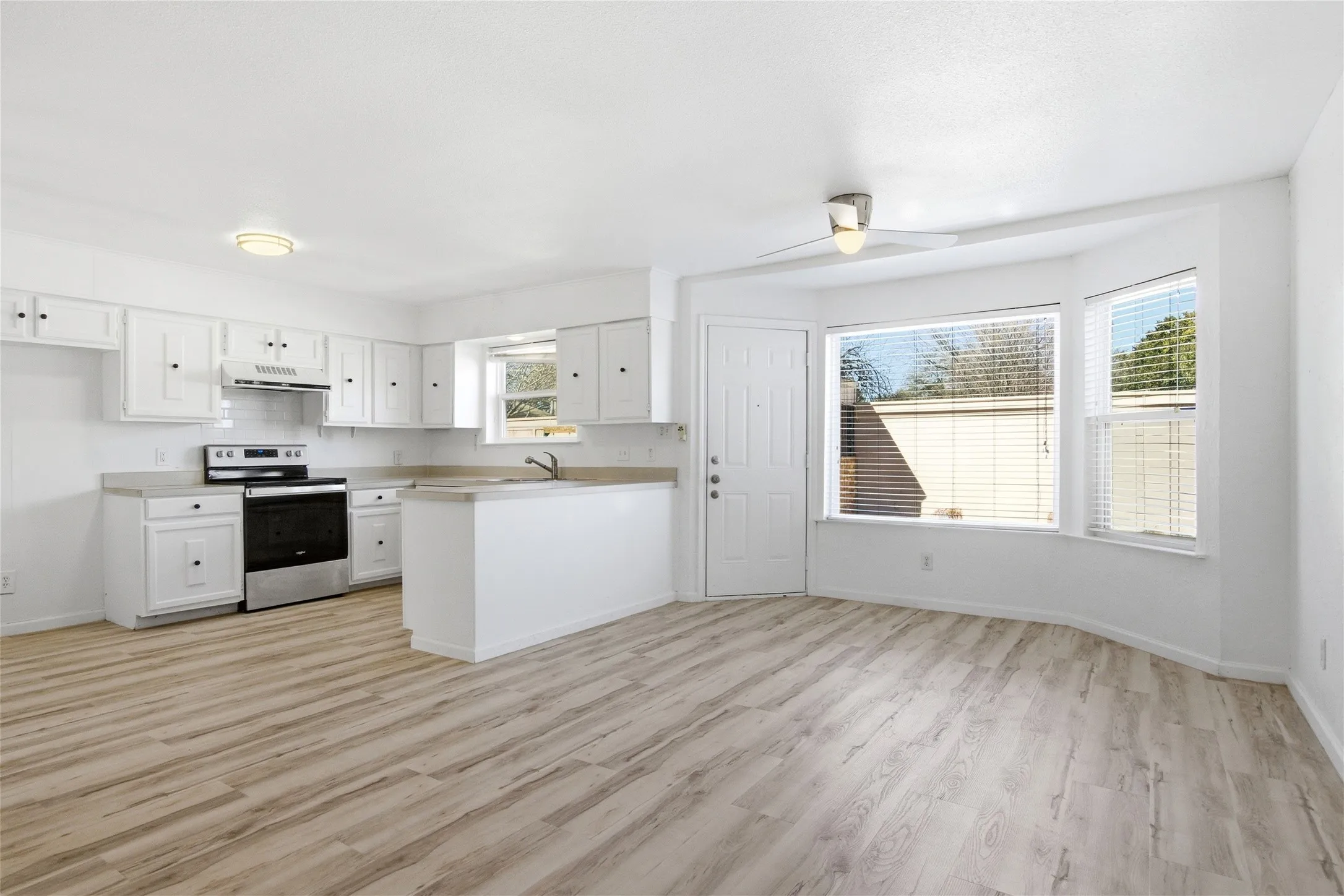 Open-concept kitchen and living area featuring wood-finish flooring, white cabinetry with black hardware, stainless steel appliances, and a bay window