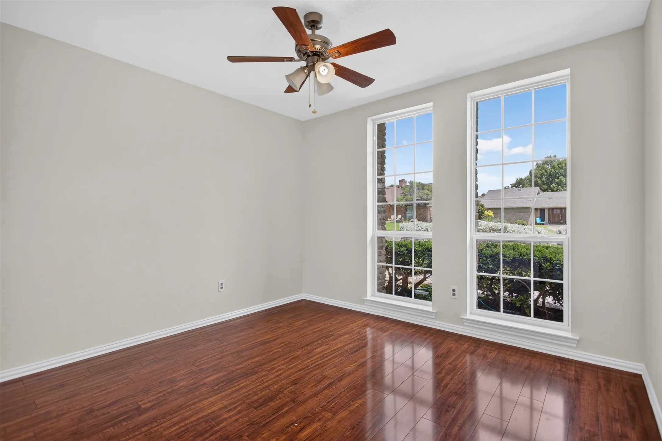Bright room featuring two tall grid-pane windows, wood-finish flooring, light neutral-toned walls, a ceiling fan with light kit, and white baseboards
