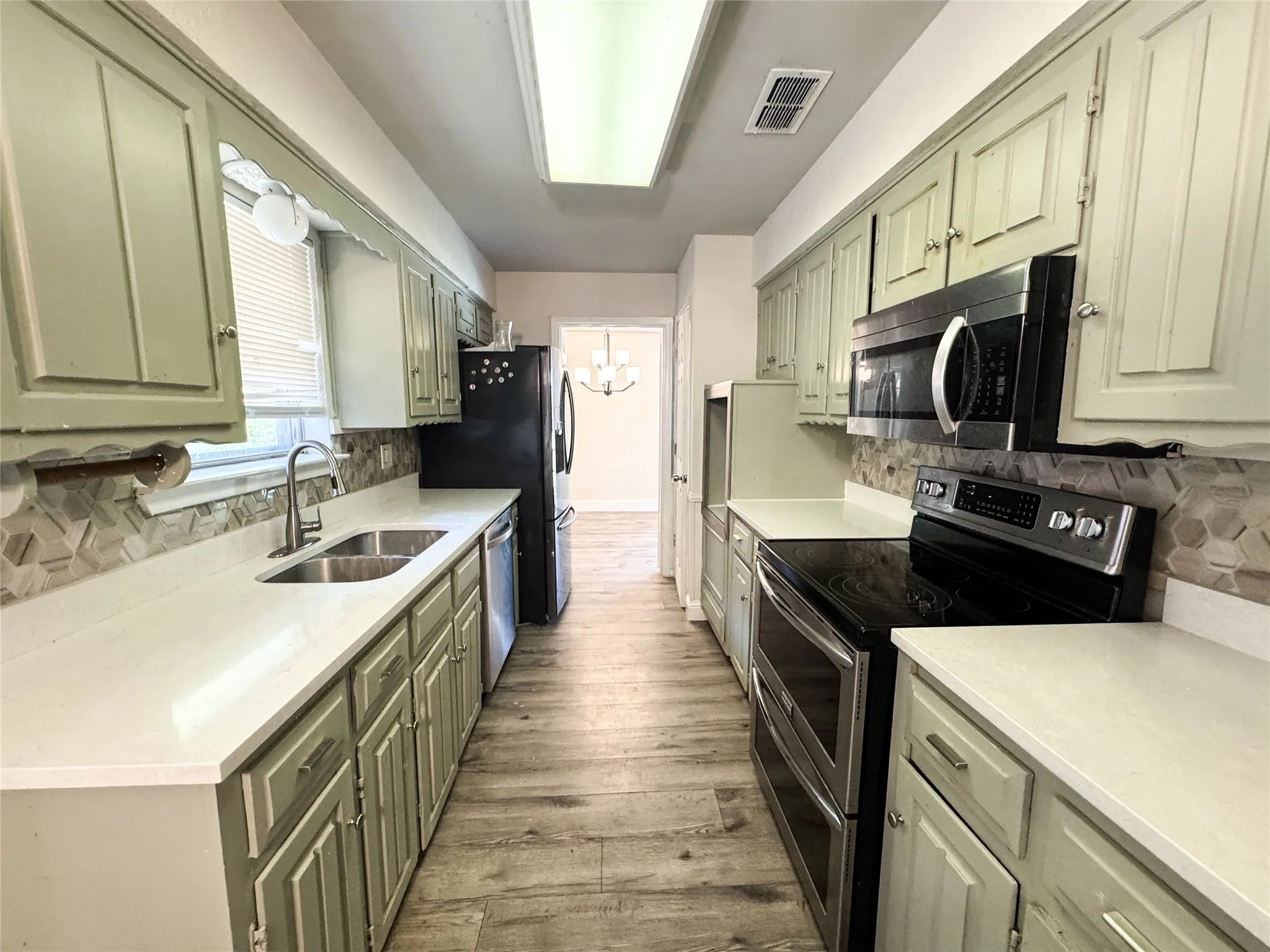 Galley kitchen featuring light green cabinetry, white countertops, stainless steel dual basin sink, black appliances, and wood-finish flooring