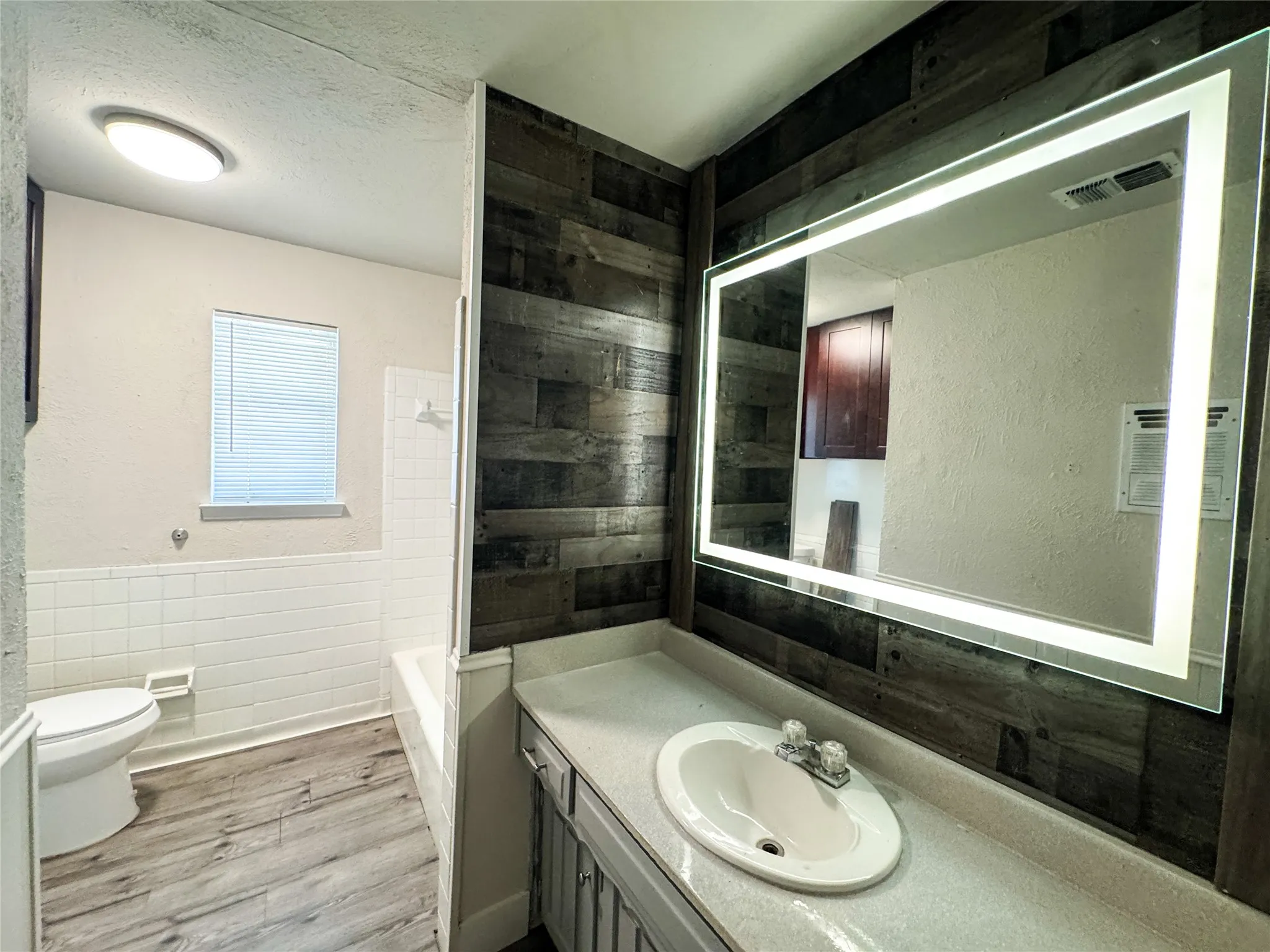 Bathroom featuring a modern backlit mirror, wood-finish accent wall, built-in vanity with integrated sink, white subway tile wainscoting, and wood-finish flooring