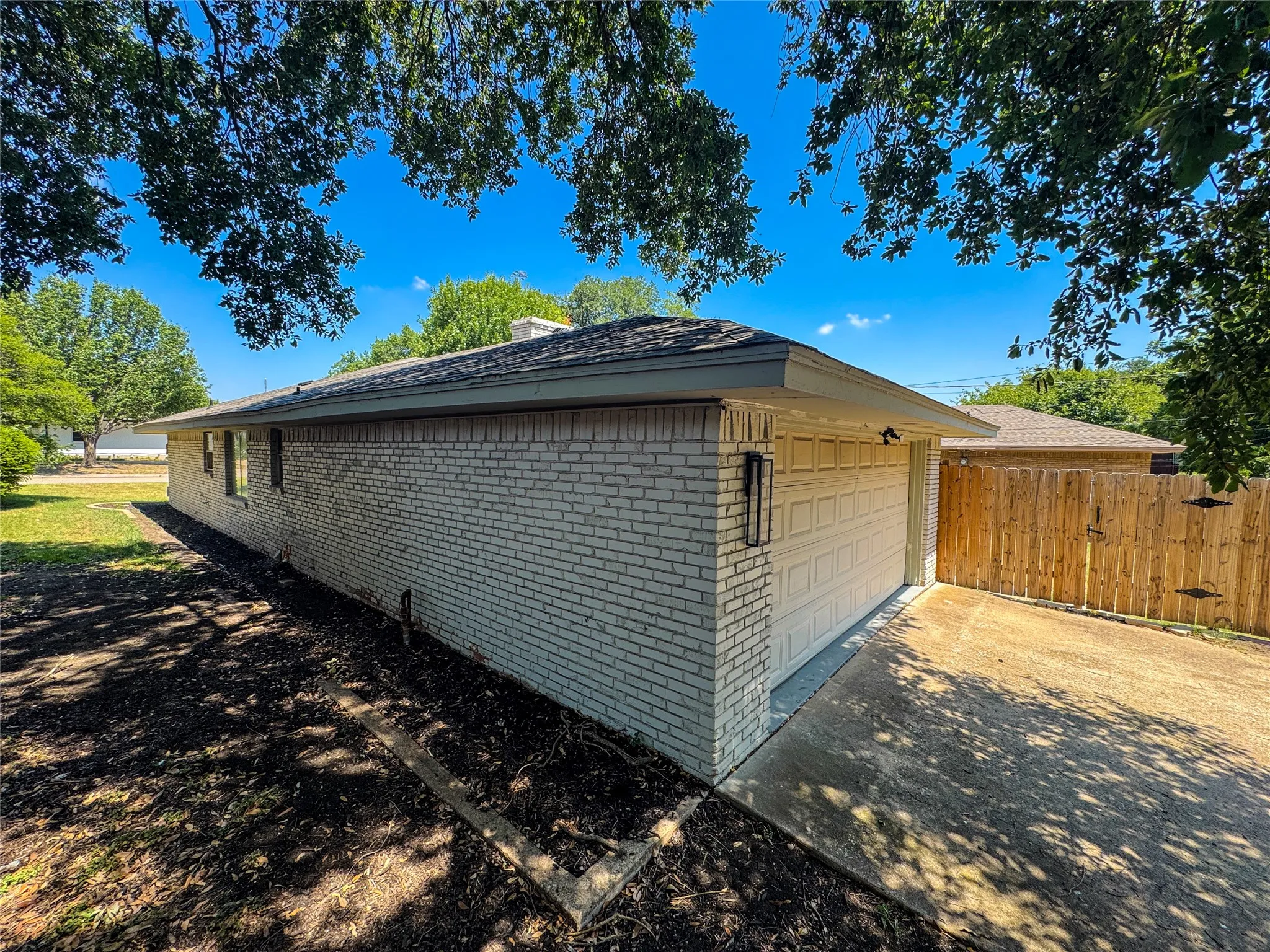 Light-colored brick exterior featuring a shingled roof, attached garage with panel door, concrete driveway, and wood privacy fencing