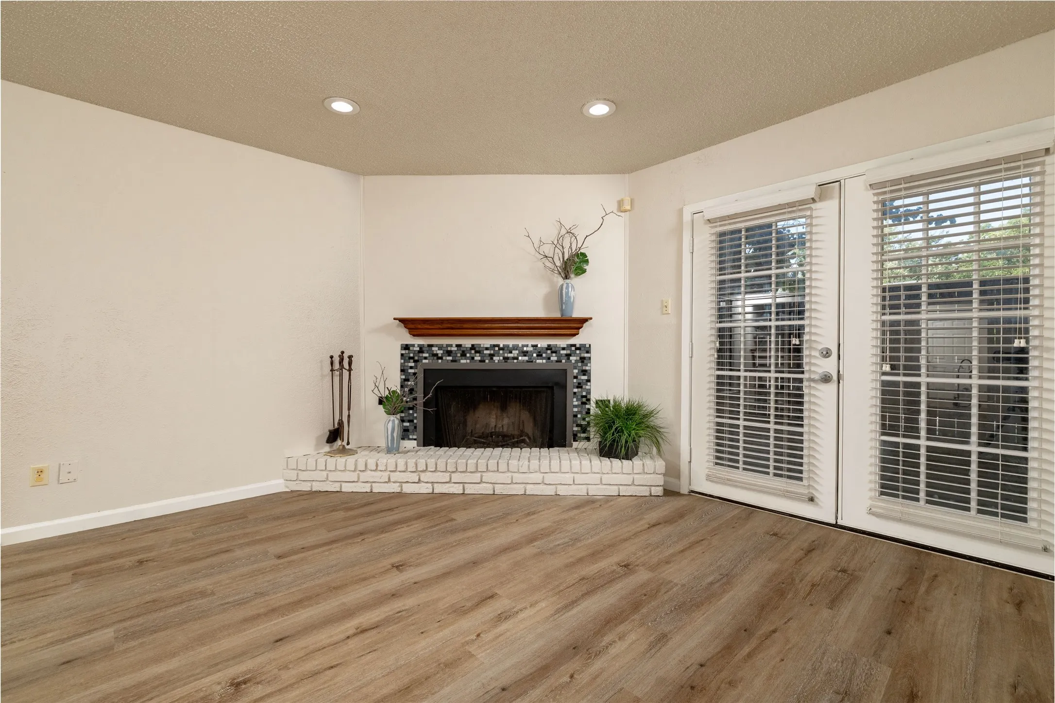 Spacious living area featuring light wood-finish flooring, a corner fireplace with a white painted brick hearth, and a mosaic tile surround