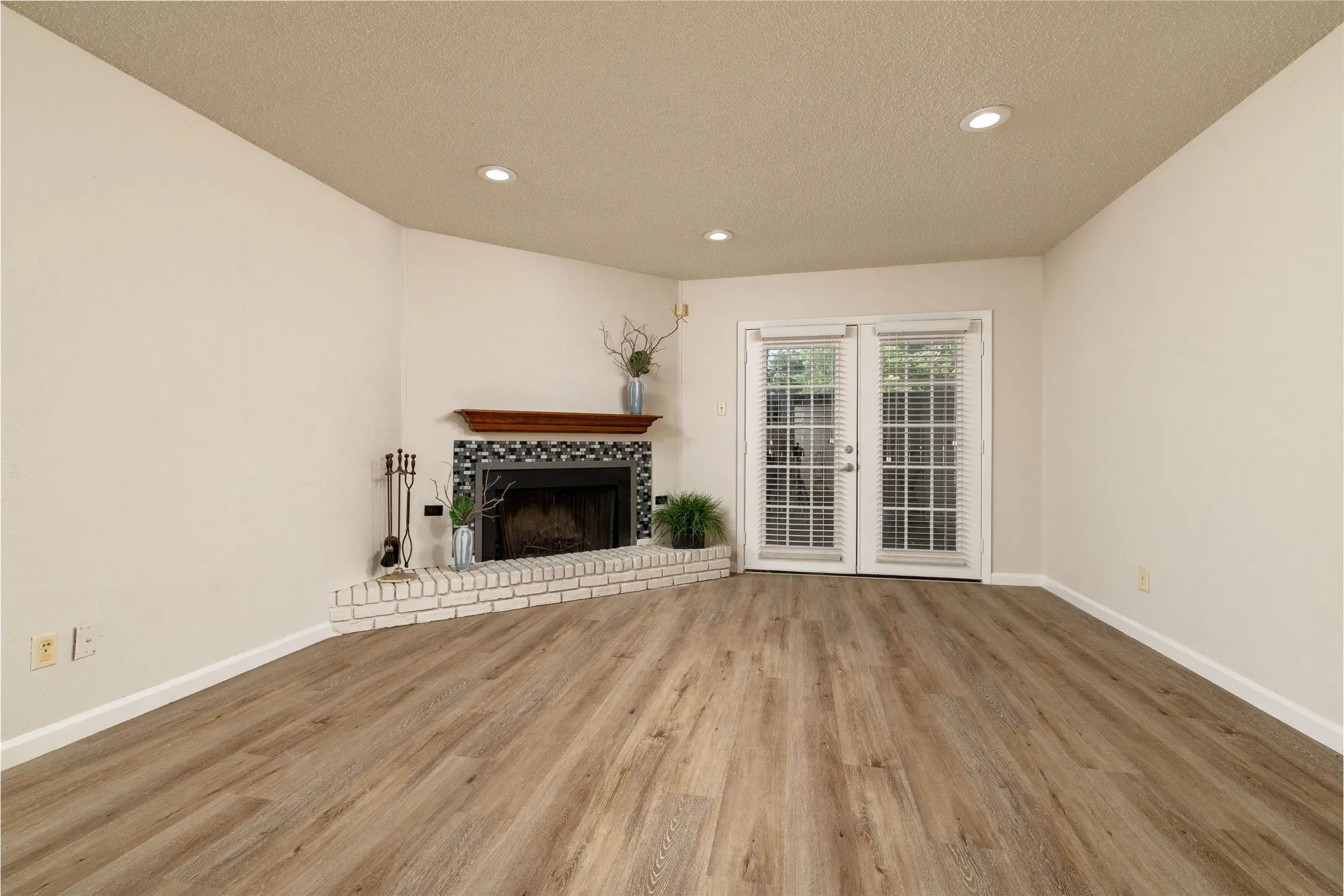 Living area featuring wood-finish flooring, a corner fireplace with a white brick hearth and decorative tile surround, a wood mantel, and recessed ceiling lighting