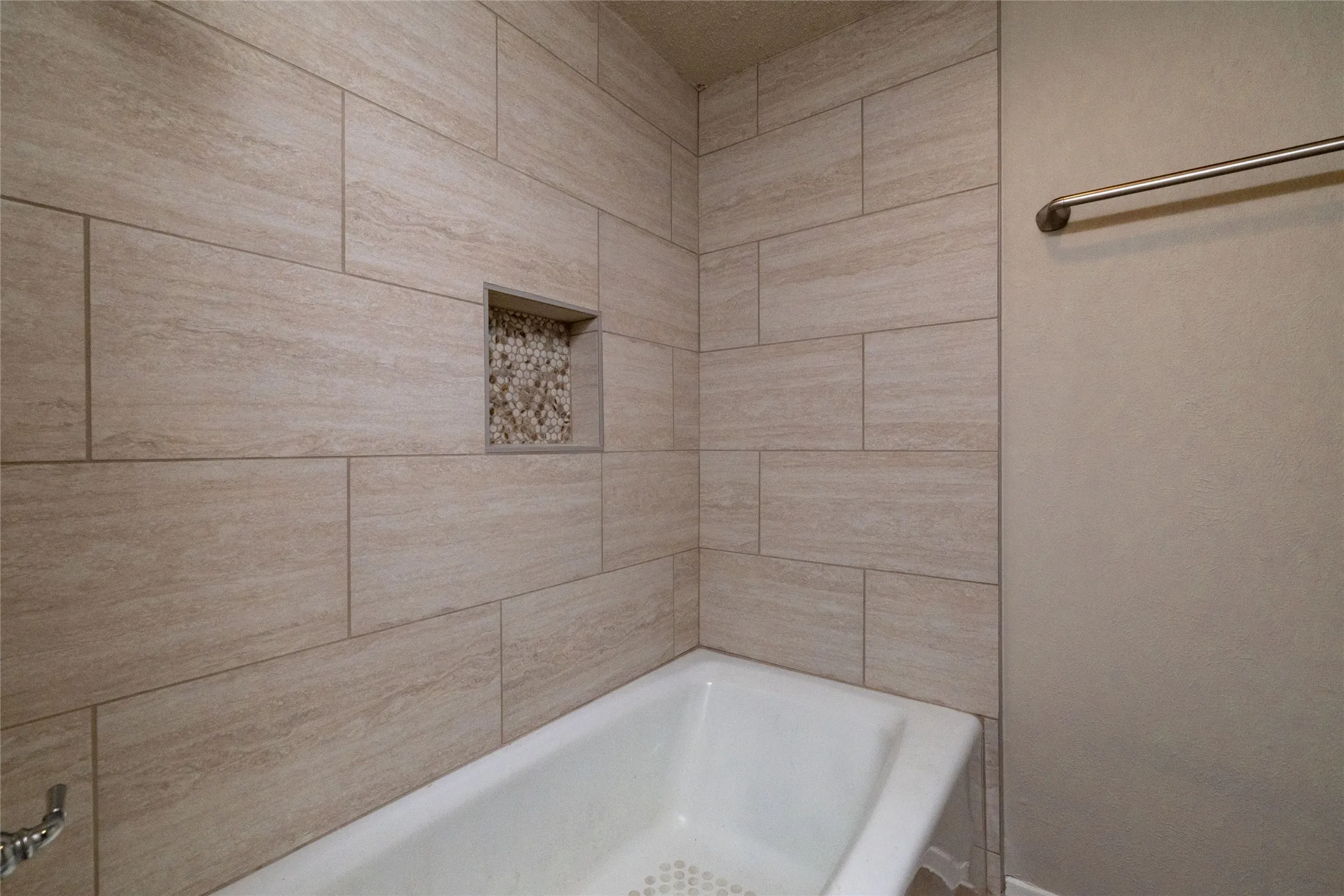 Porcelain tile shower surround with a built-in niche featuring pebble tile accent, a white bathtub, and a chrome grab bar