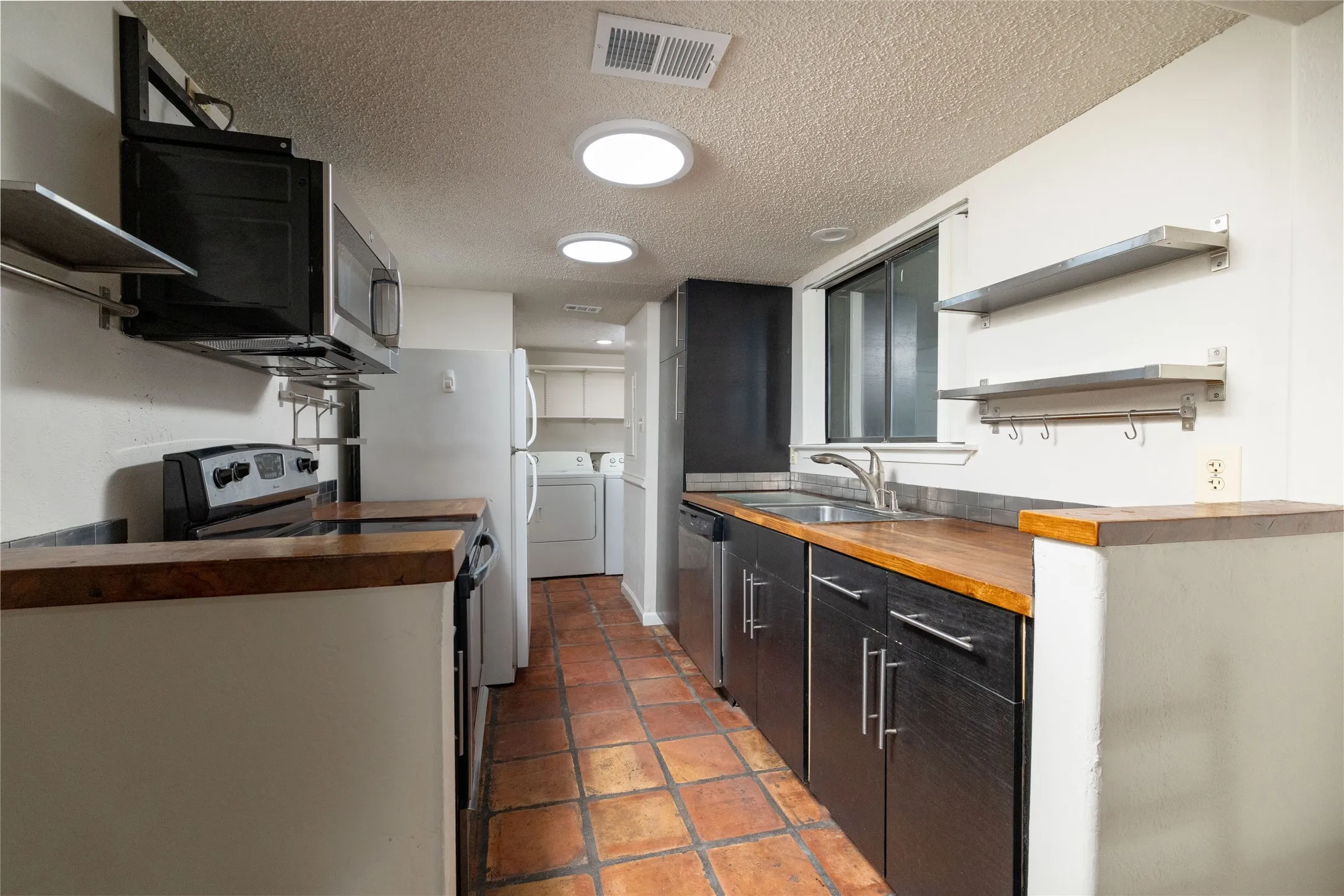 Galley kitchen featuring wood-finish countertops, dark cabinetry, and terra cotta tile flooring