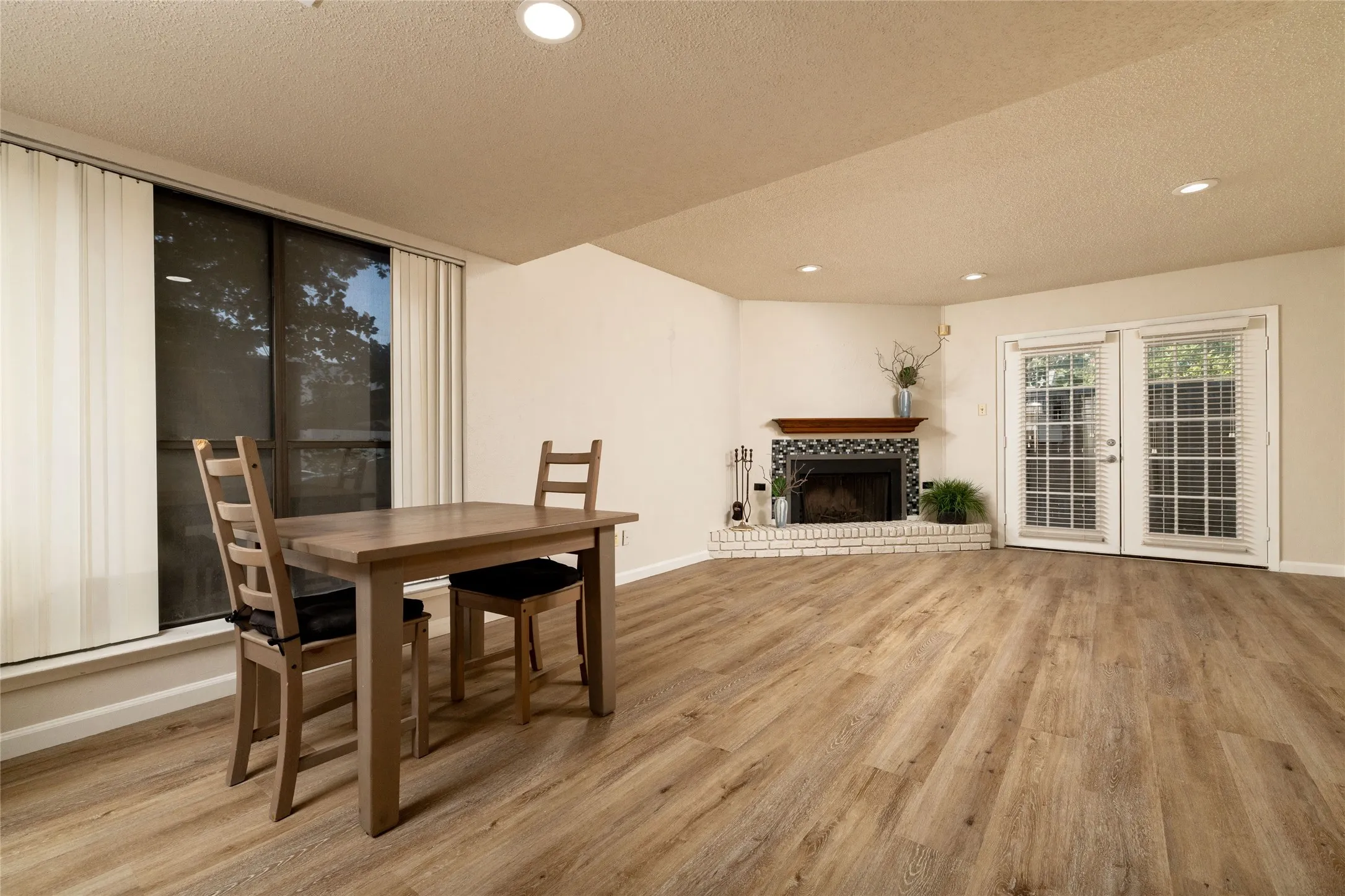 Spacious open-concept room featuring wood-finish flooring, recessed lighting, and a corner fireplace with a white brick hearth and decorative tile surround