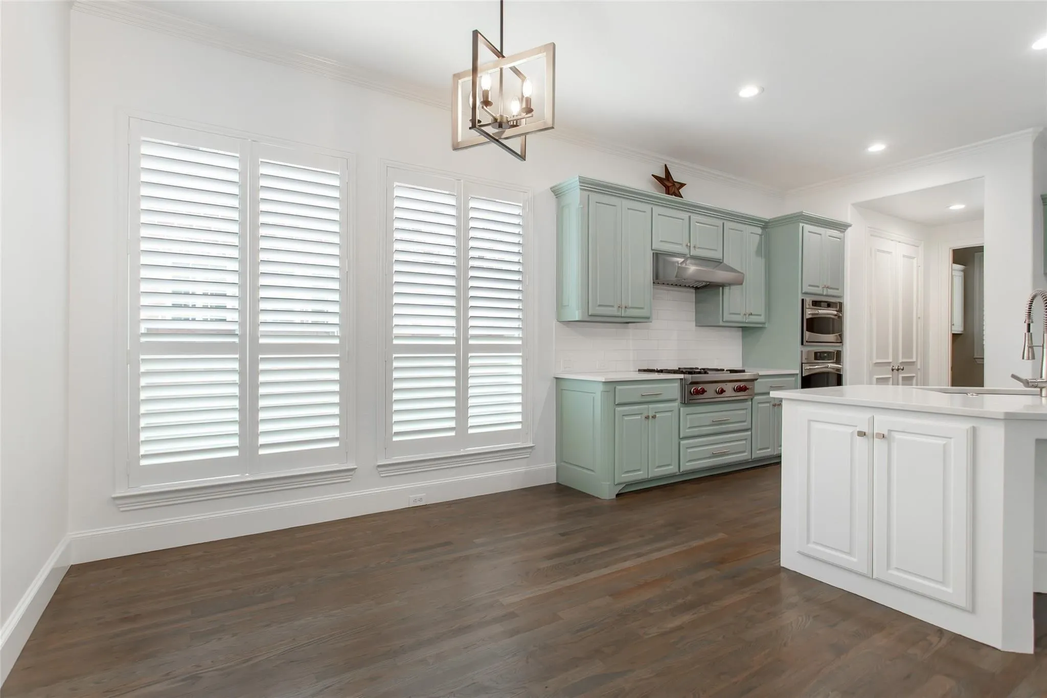 Expansive kitchen and dining area featuring wide plank wood-finish flooring, three large windows with plantation shutters, custom cabinetry, a built-in range, and a kitchen island with storage