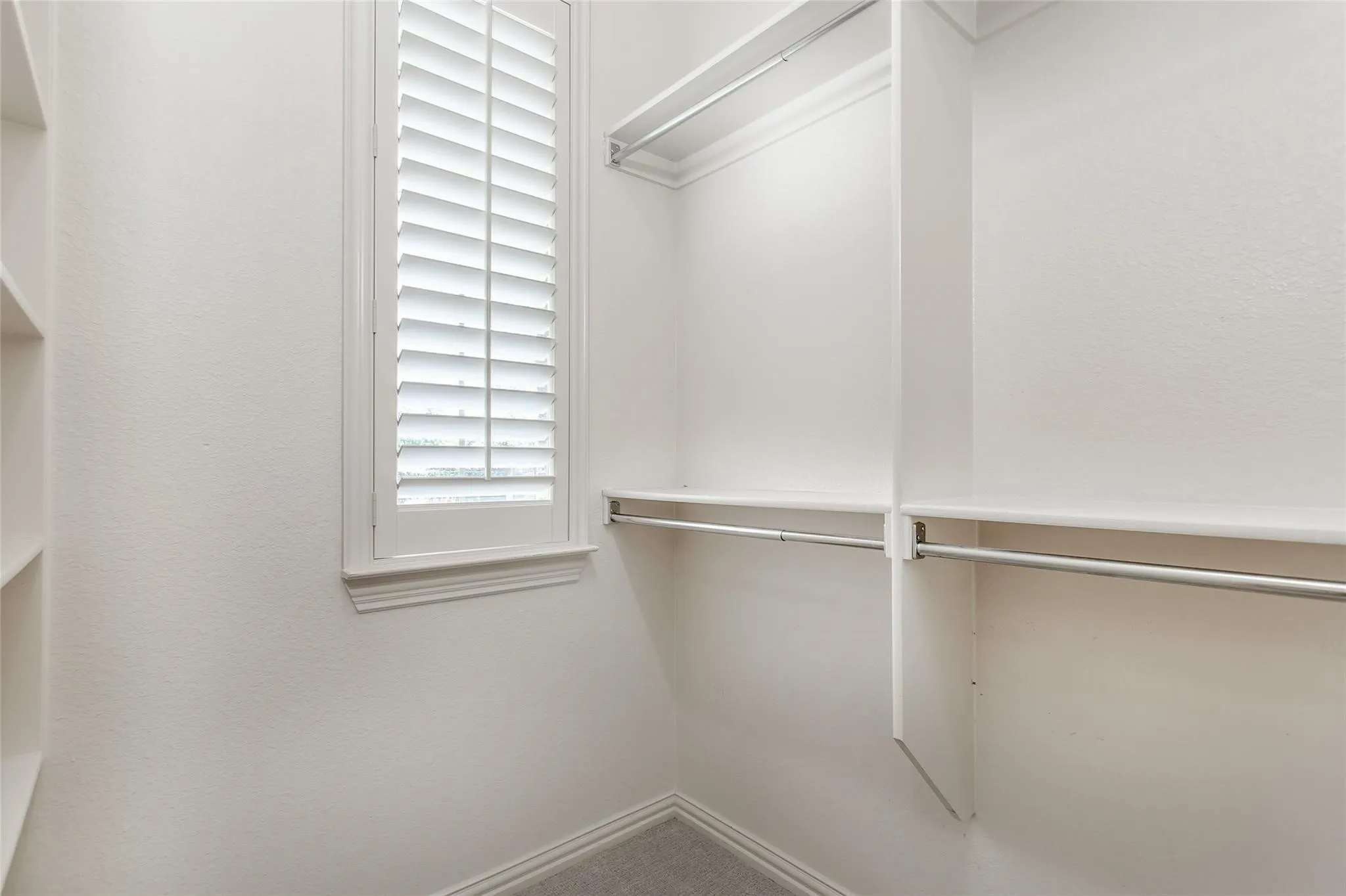 Built-in closet system featuring multiple shelves and hanging rods, a window with plantation shutters, and light-colored carpeting
