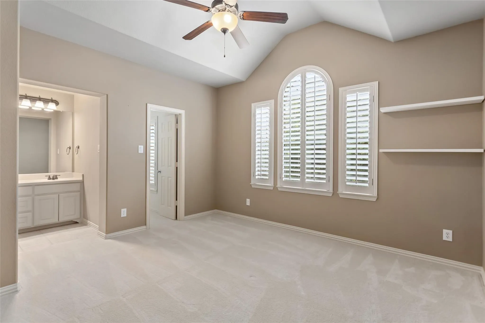Vaulted ceiling with a ceiling fan, three windows with plantation shutters, light-toned carpeting, built-in shelving, and an en-suite vanity area with a built-in sink and light fixture
