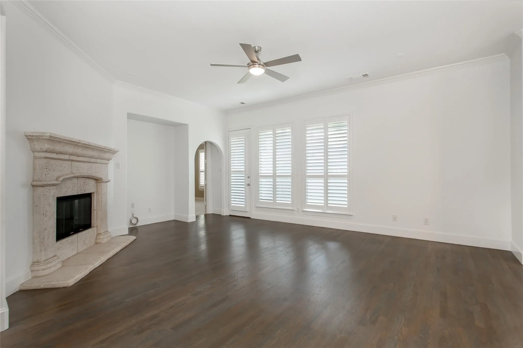 Spacious room featuring dark wood-finish flooring, a stone fireplace with a hearth, multiple windows with white plantation shutters, an arched doorway, and a ceiling fan