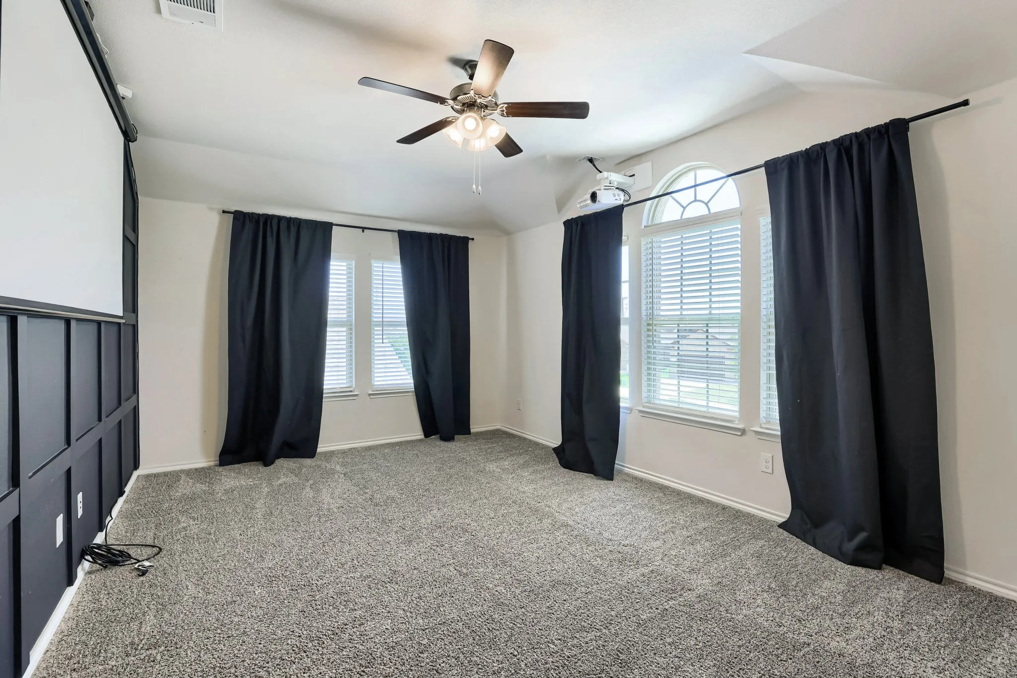 Projector screen with paneling, vaulted ceiling, ceiling fan with integrated lighting, arched window, and gray carpet