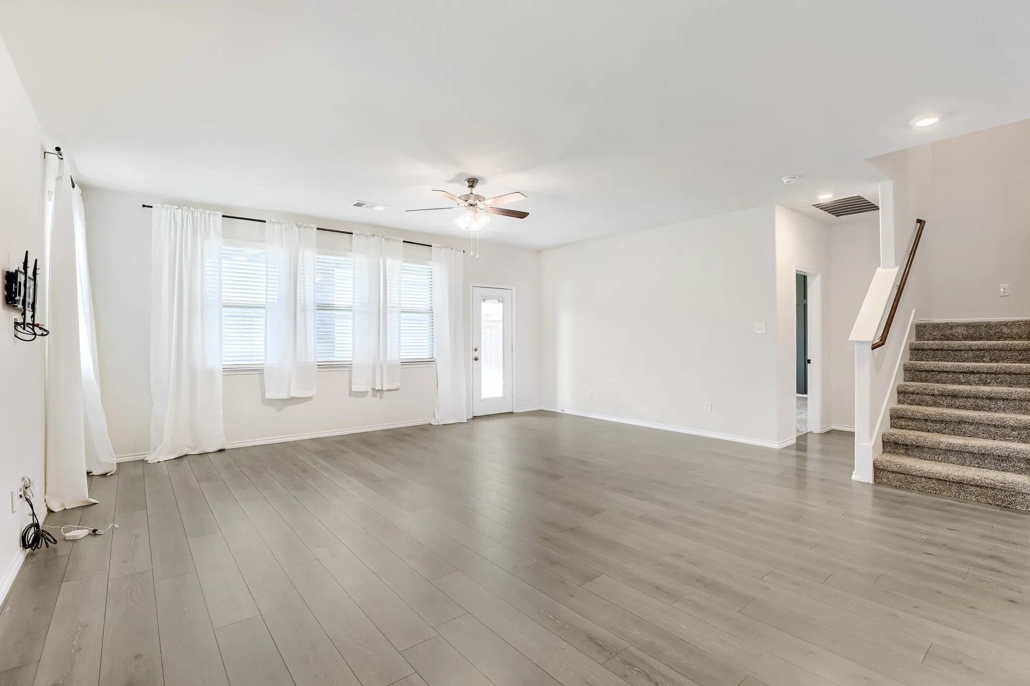 Spacious room featuring wood-finish flooring, a ceiling fan, multiple windows with blinds, a glass-paneled door, and a carpeted staircase with a wood handrail