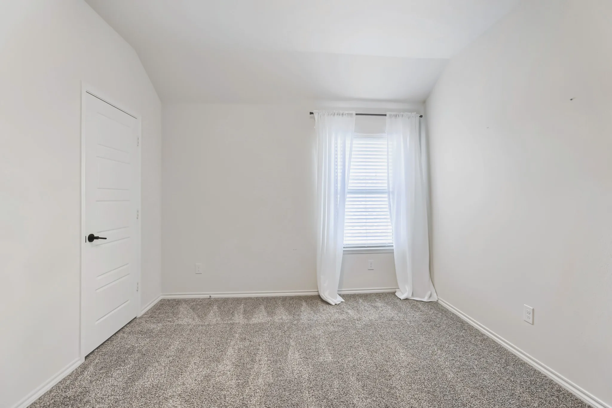 Neutral paint tones, vaulted ceiling, grey carpet, window with blinds, and a modern paneled door with black hardware