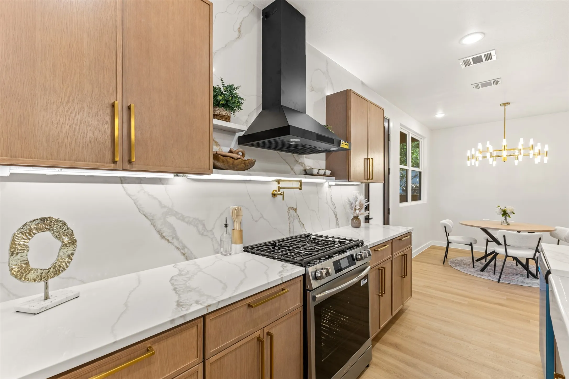Contemporary kitchen featuring light wood cabinetry with gold-finish hardware, a gas range with a black range hood, and white countertops with a matching backsplash