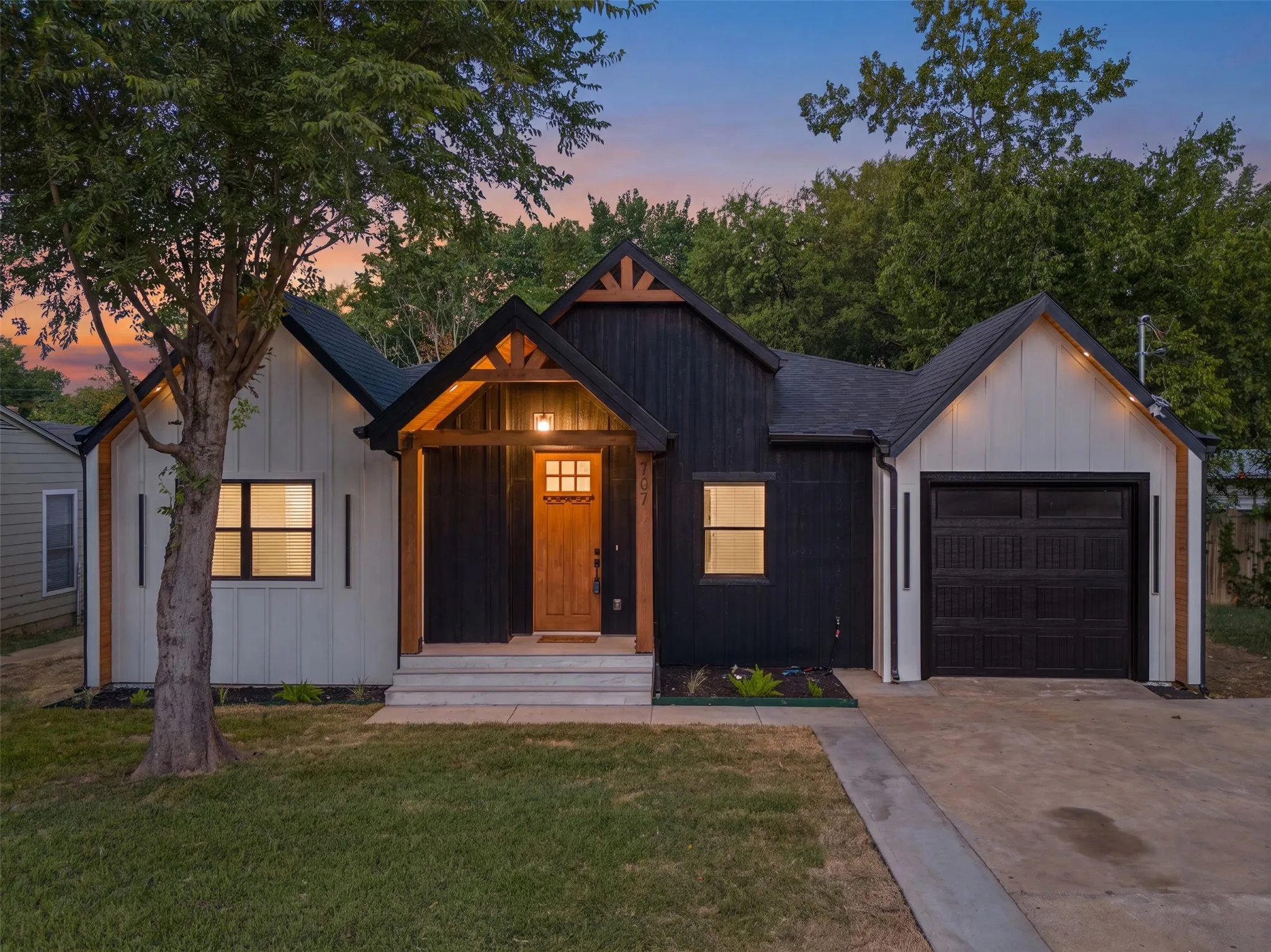 Modern farmhouse-style exterior featuring a striking black and white color palette, a covered entry with exposed wood beams, a natural wood front door with grid-patterned glass, a single-car garage with a paneled door, and a concrete driveway
