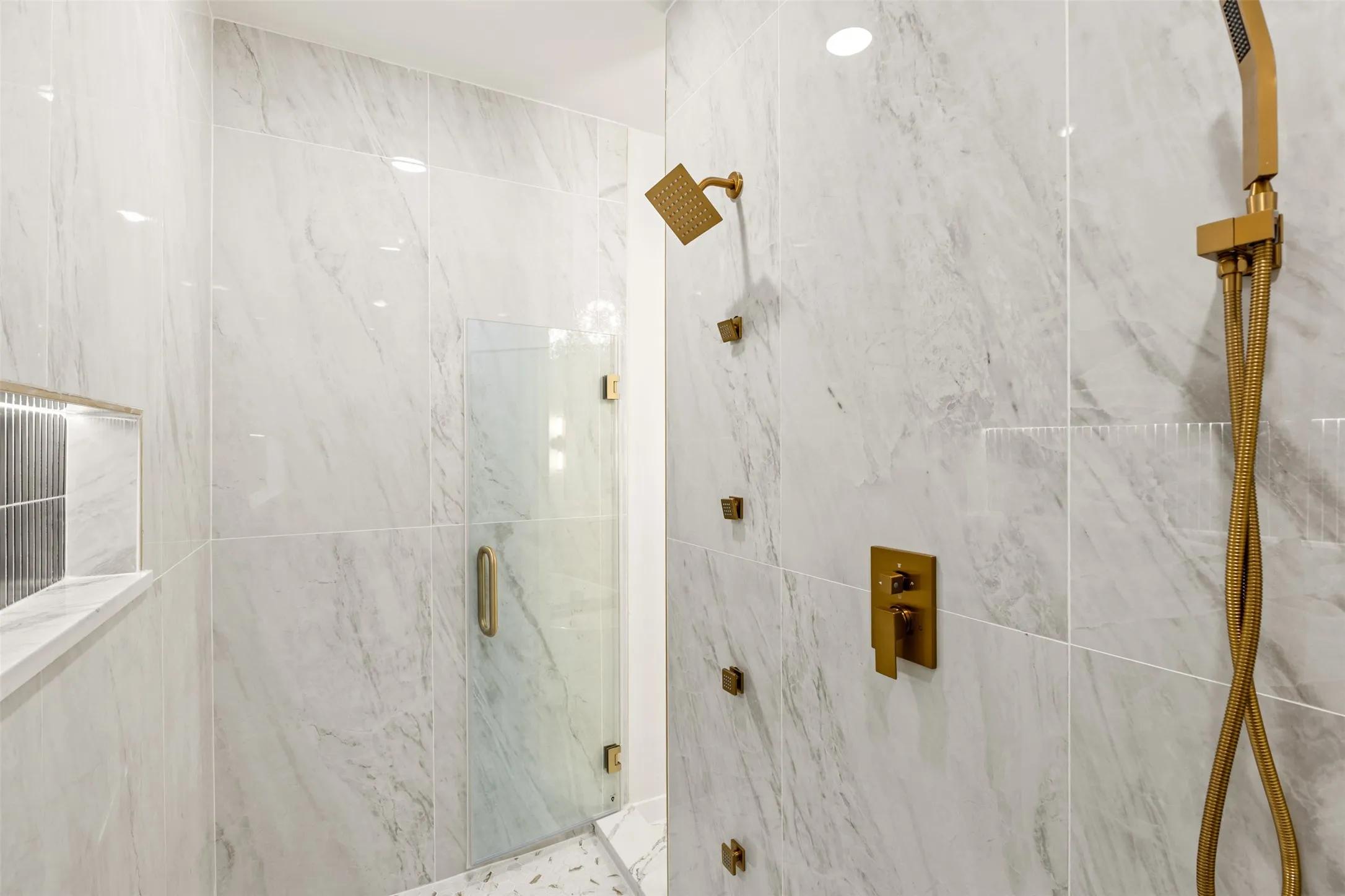 Walk-in shower featuring large format marble-look tile, gold-tone fixtures, a built-in shower niche with accent tile, and a frameless glass enclosure
