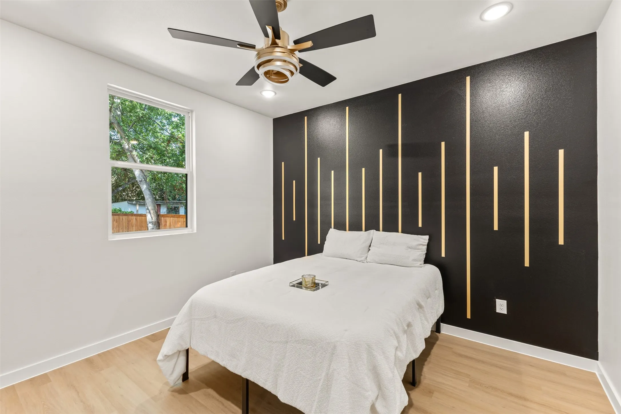 Bedroom featuring an accent wall with vertical wood paneling, light wood-finish flooring, a white trim baseboard, a window, and a ceiling fan with integrated lighting